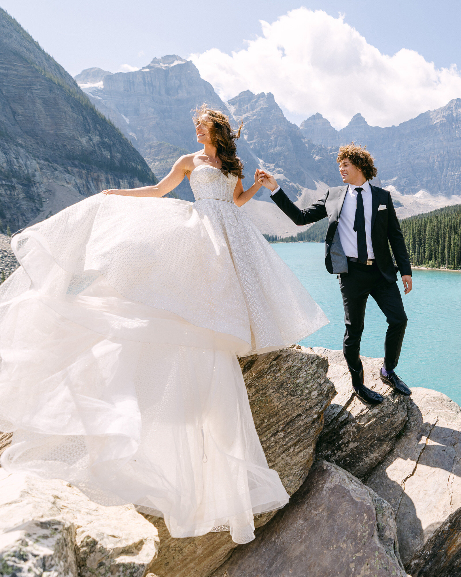 A breathtaking moment as the bride and groom hold hands, surrounded by the iconic Valley of the Ten Peaks in Moraine Lake, Banff National Park. With the bride elegantly placing her dress, this stunning photograph captures the romance, grandeur, and timeless beauty of their unforgettable wedding day in the Canadian Rockies.
