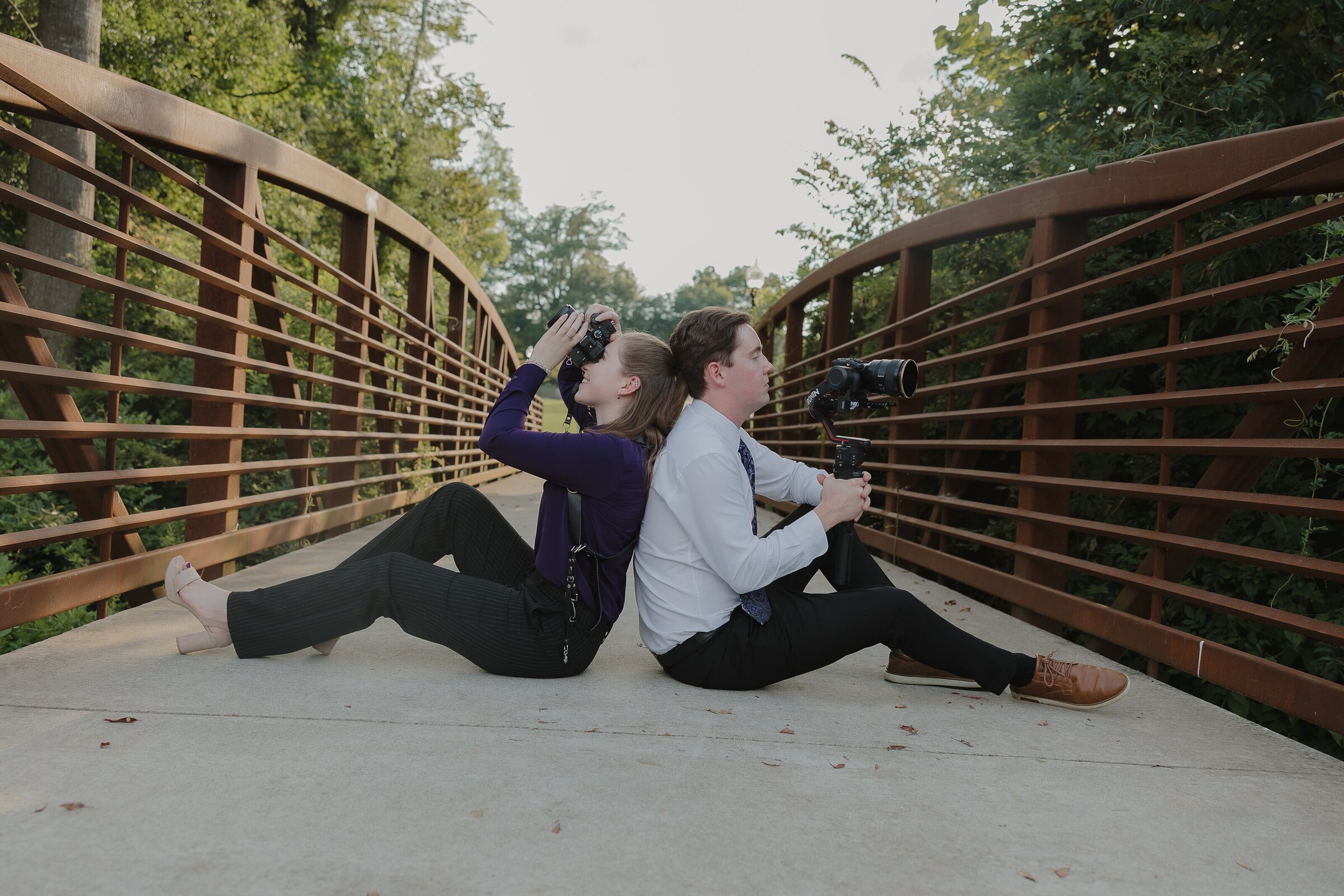 Wedding photo and video team sitting back-to-back on the Depot Park bridge in Kennesaw with their cameras.