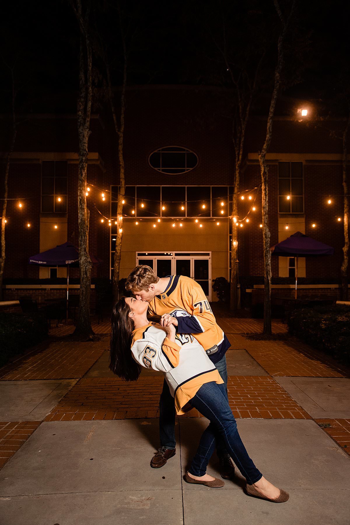 Couple wearing their Nashville predator jerseys in a courtyard at night with bistro lights in the background