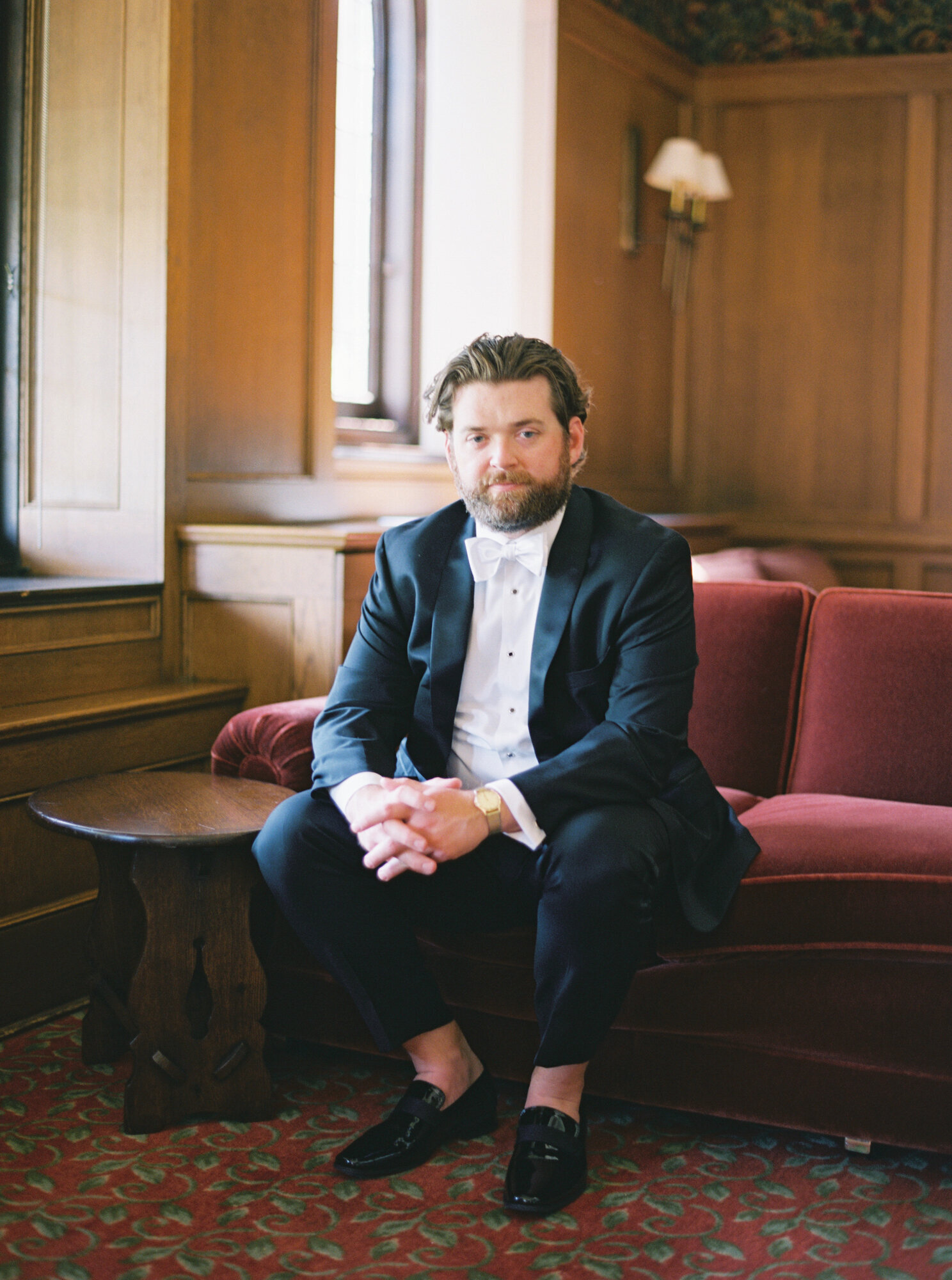 portrait of groom in black suit on red velvet couch in historic venue