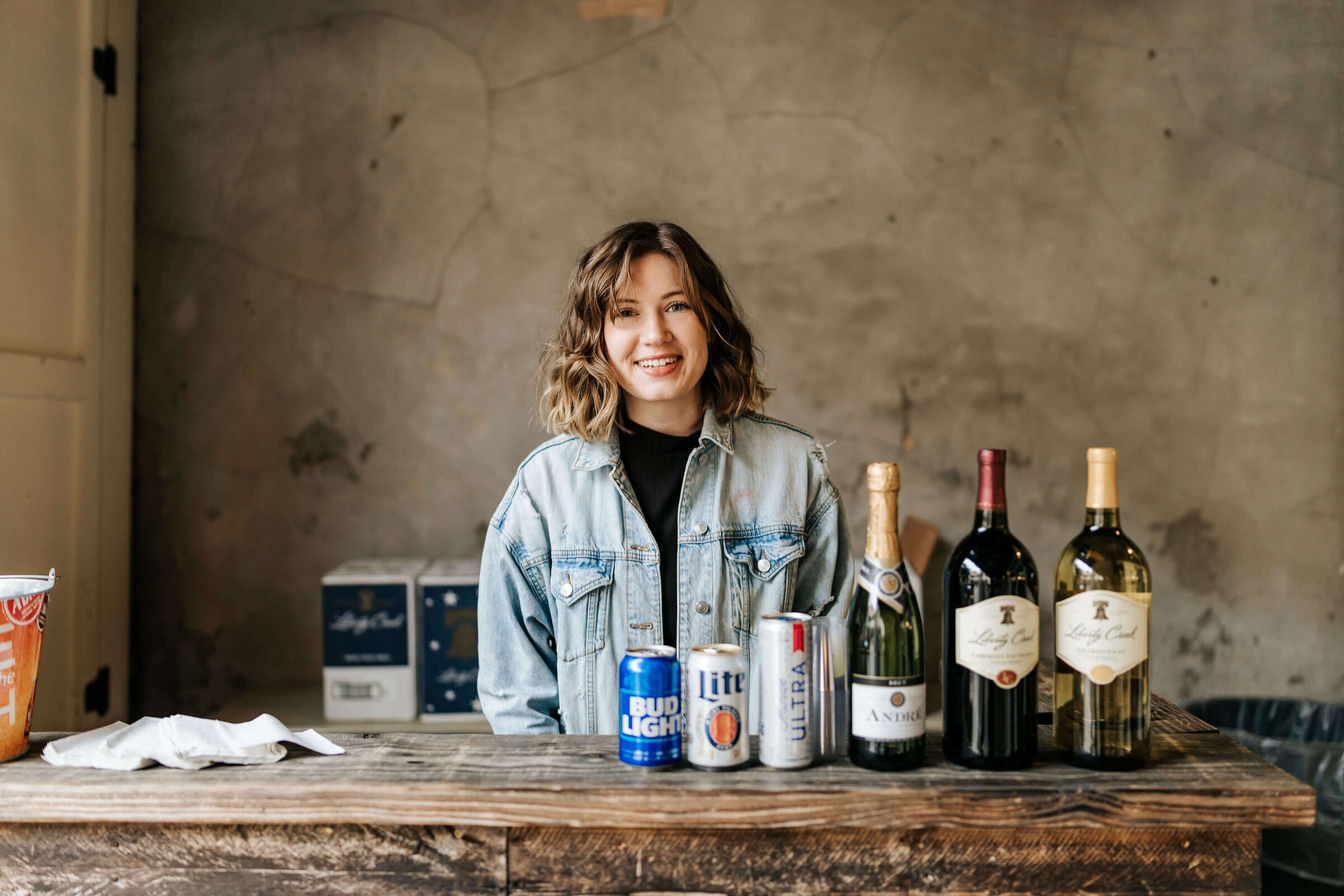 A woman with wavy hair wearing a denim jacket stands behind a wooden counter with various bottles of displayed in front of her.