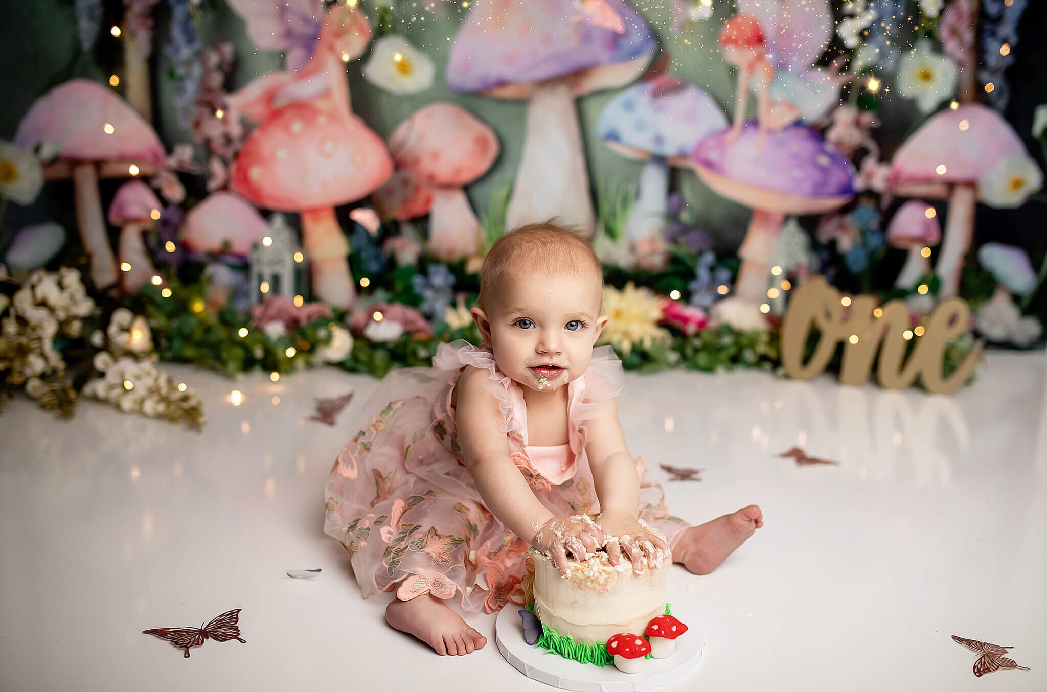 One year old cake smash with 1 year old girl sitting in front of a fairy tale background with mushrooms and fairy lights while smooshing a cake with her hands