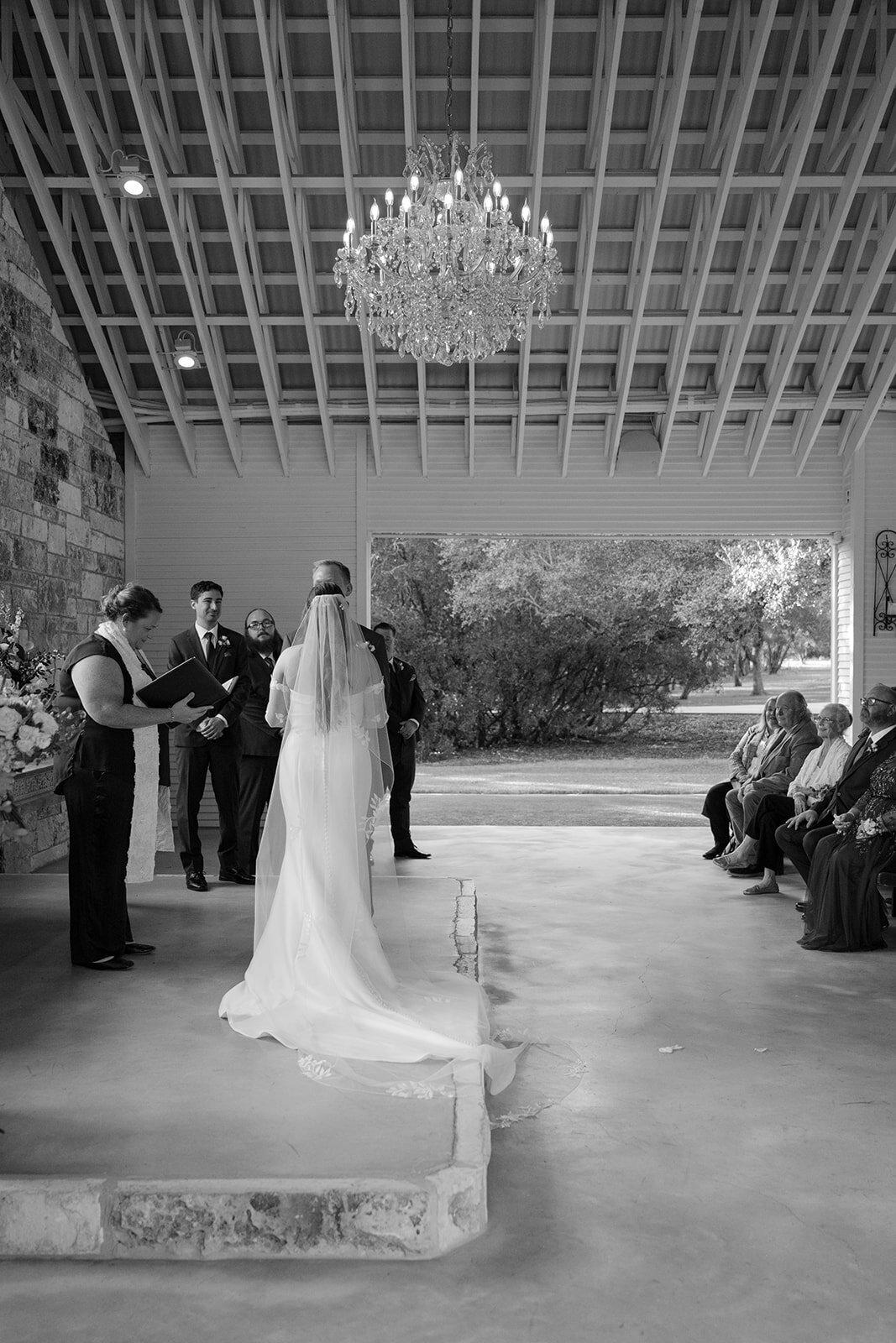 Black and white photo of a bride and groom holding hands at the front of the altar at an elegant open air wedding ceremony at The Chandelier of Gruene Texas, surrounded by guests, photographed by Erin Marcin.