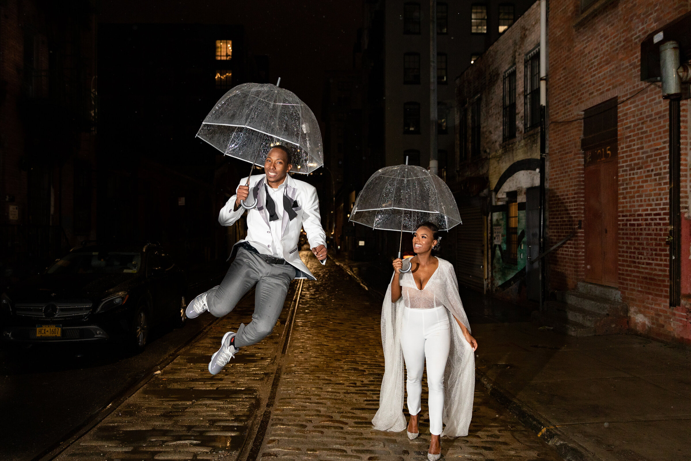 A couple walking along a sidewalk while holding umbrella while one of them jumps.