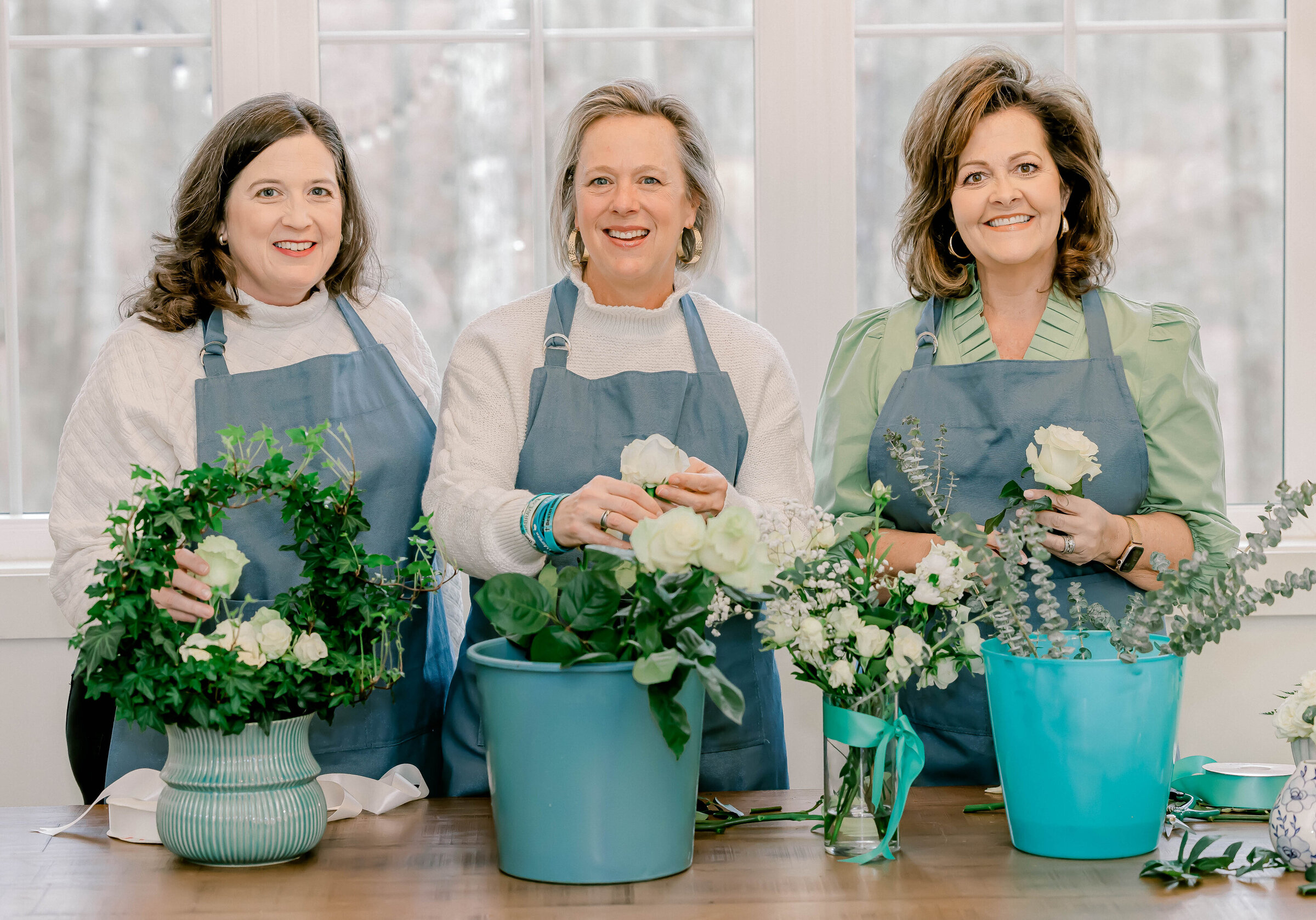 dark haired woman holding flowers on the left, in the center blonde woman holding flowers, and on the right dark haired woman holding flowers
