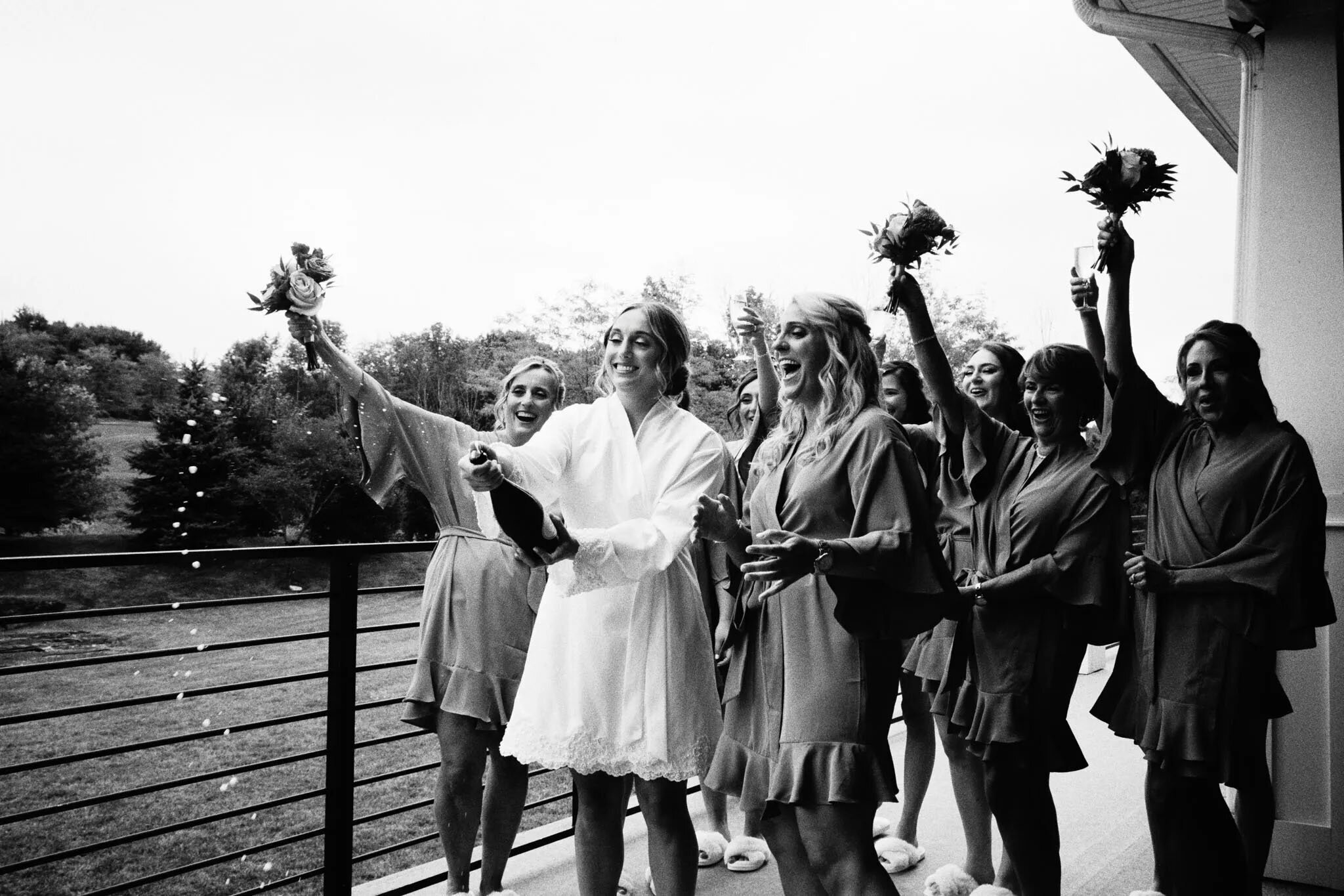 A bride in a white robe pops a champagne bottle on a balcony, surrounded by bridesmaids in matching robes holding bouquets, all smiling and celebrating—captured beautifully by an NJ wedding photographer with trees and greenery in the background.
