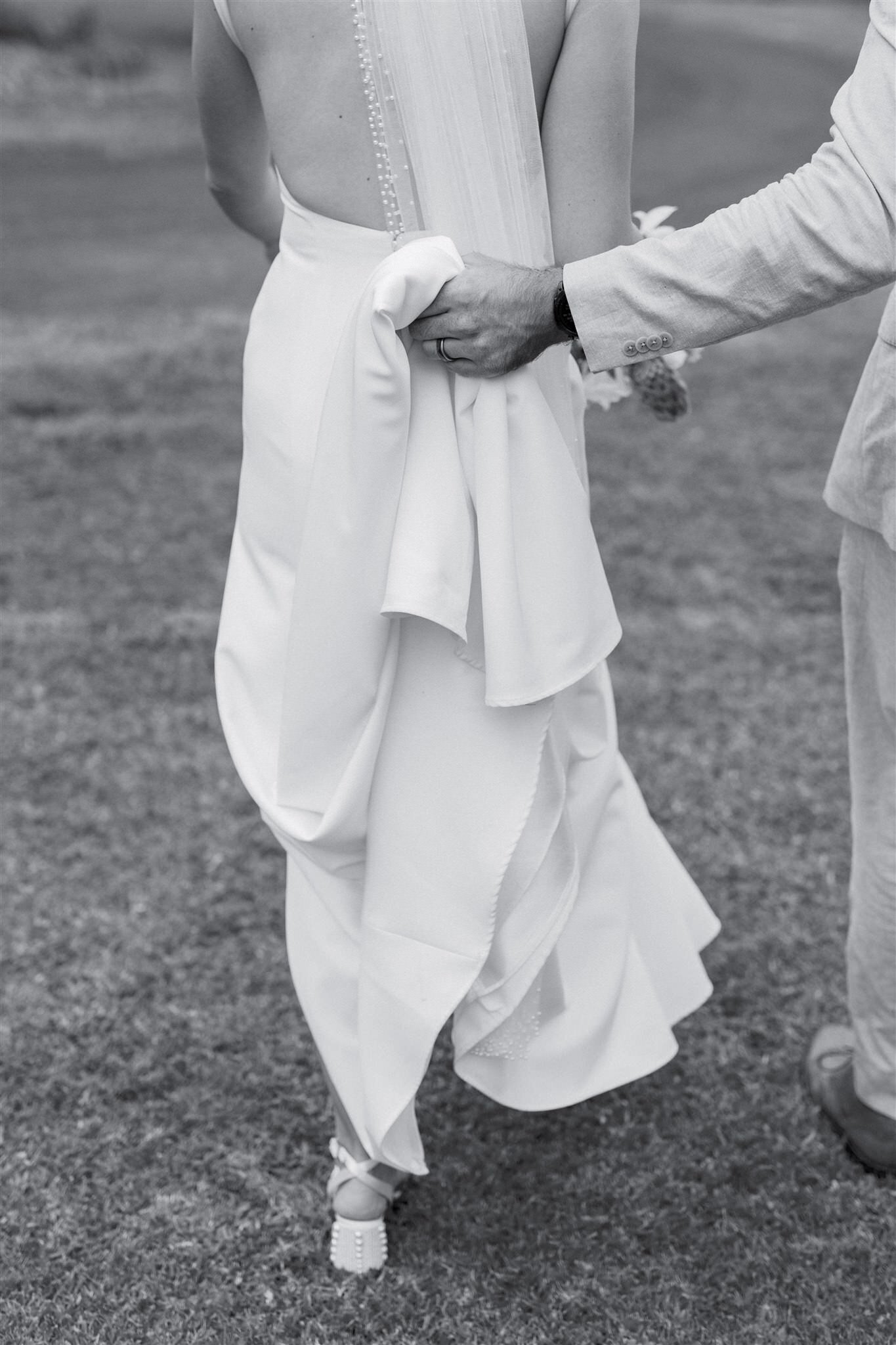 A person holding the train of their partner's wedding dress as they walk 