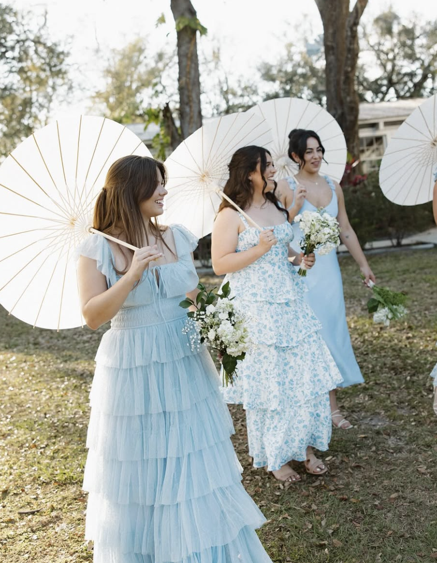 Bridesmaids in pastel blue dresses holding white parasols and bouquets, smiling as they walk the grounds of Uliana Estate in Lakeland, Florida