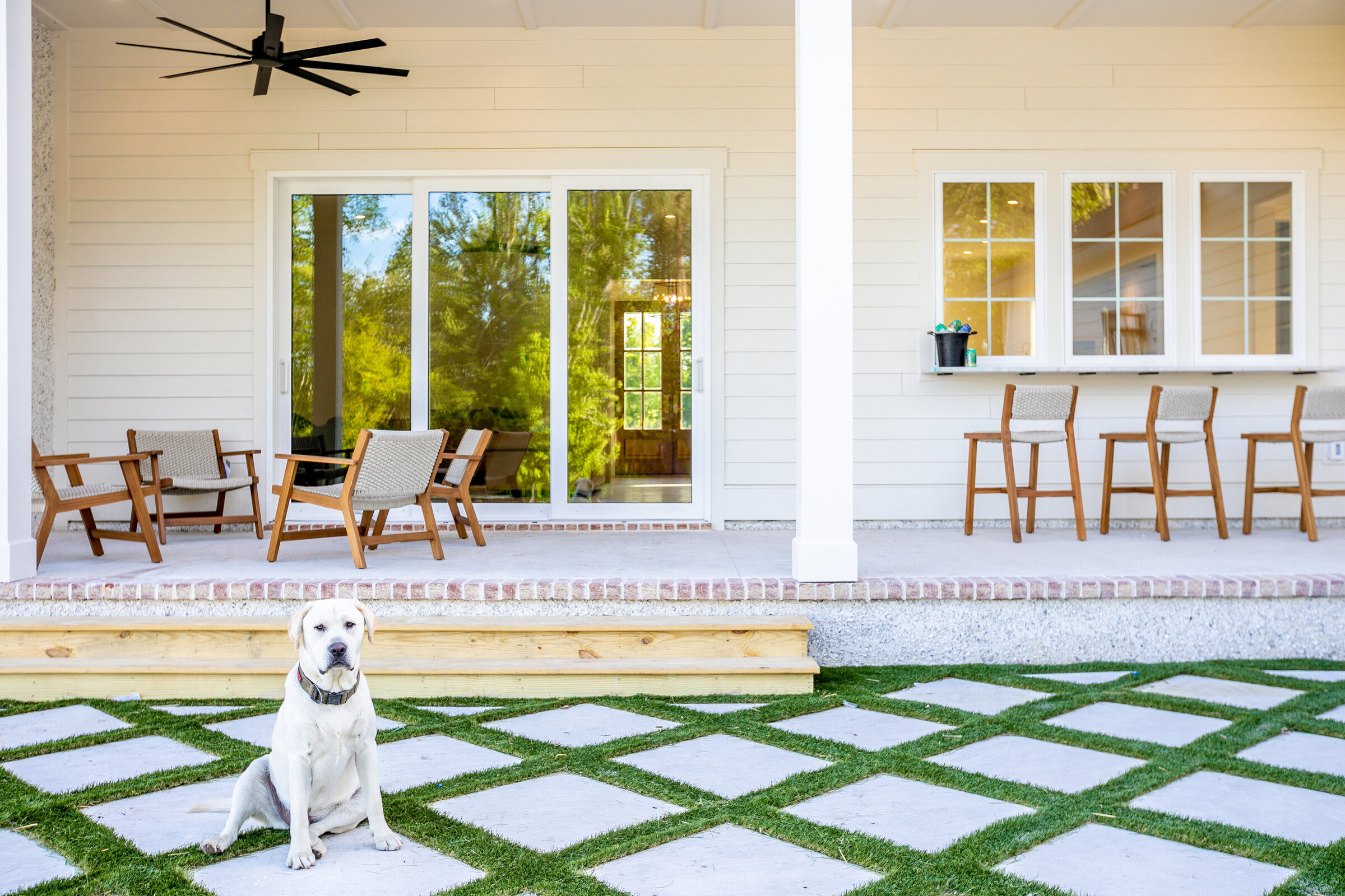 An exquisite outdoor living space featuring a bar with a window open to the kitchen, and a cozy sitting area. The pavers, designed with turf between them, create a luxurious checkered pattern that enhances the sophisticated ambiance of this long shore home.