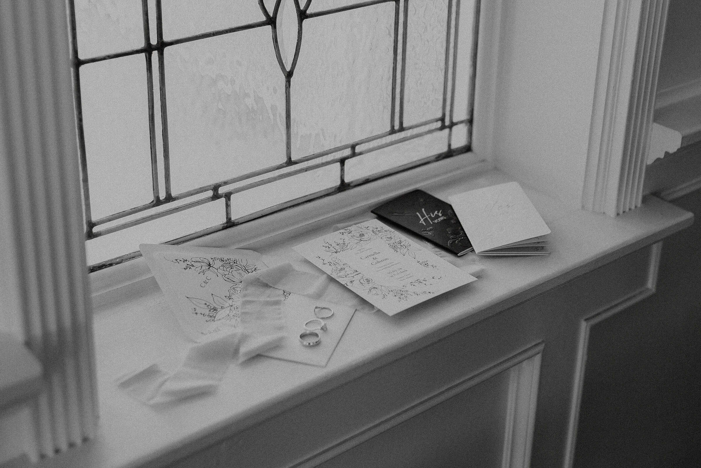 black and white photo  of an invitation suite, voew books, wedding rings, and ribbon laying on a windowsill at lindsey grove wedding venue in mesa arizona