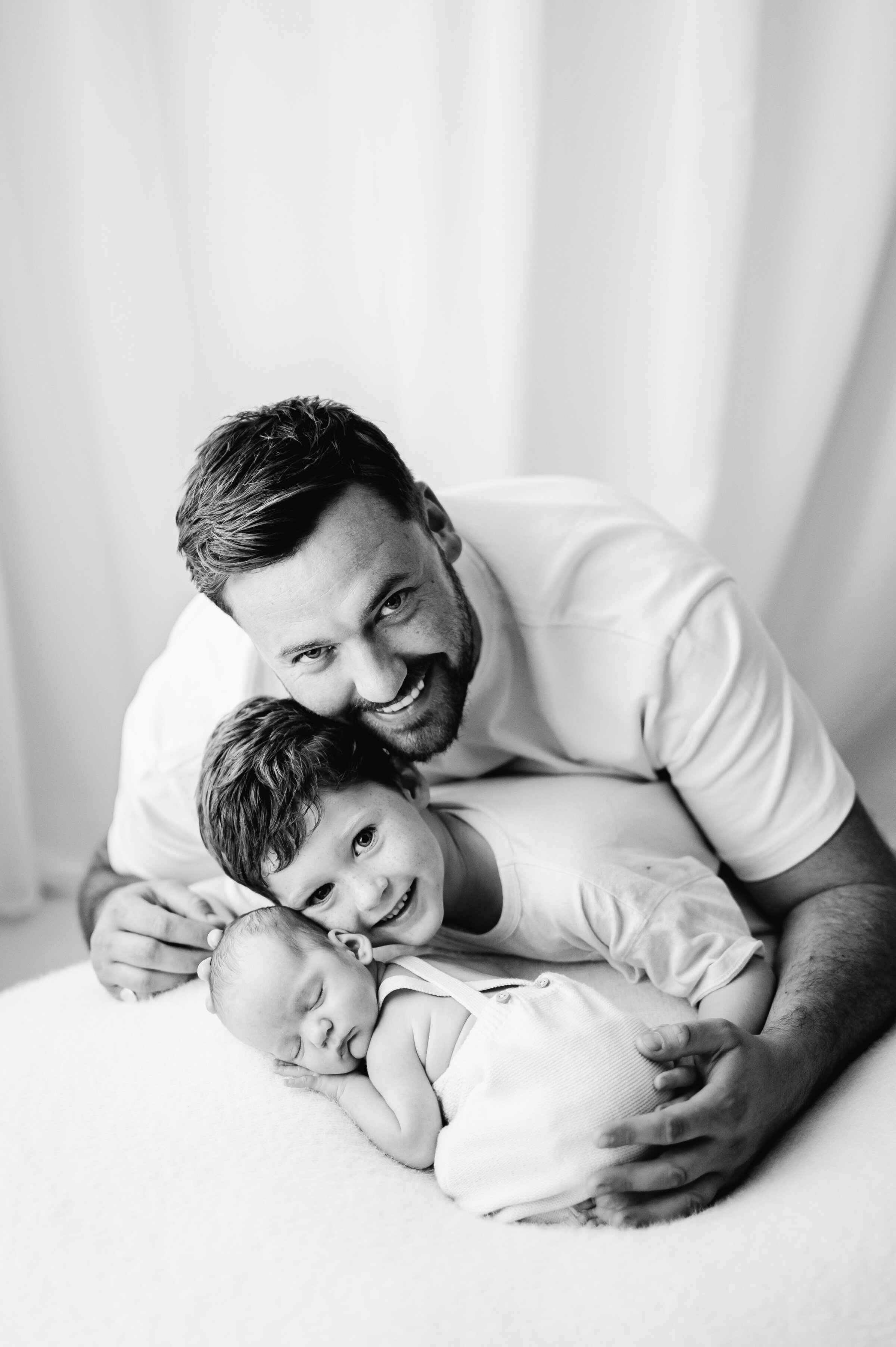 A dad and his son leaning over the top of a newborn boy sleeping on a white blanket at Billingshurst newborn photoshoot