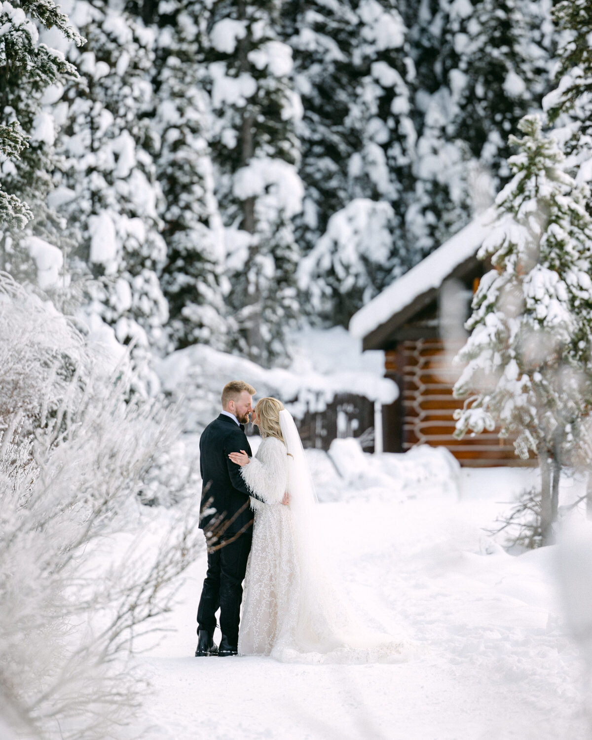 A magical winter elopement photograph as the bride and groom share a kiss while walking through the snow at Chateau Lake Louise. With the charming boathouse and frozen lake in the background, this intimate scene captures the beauty and romance of a perfect winter’s day in Banff National Park.