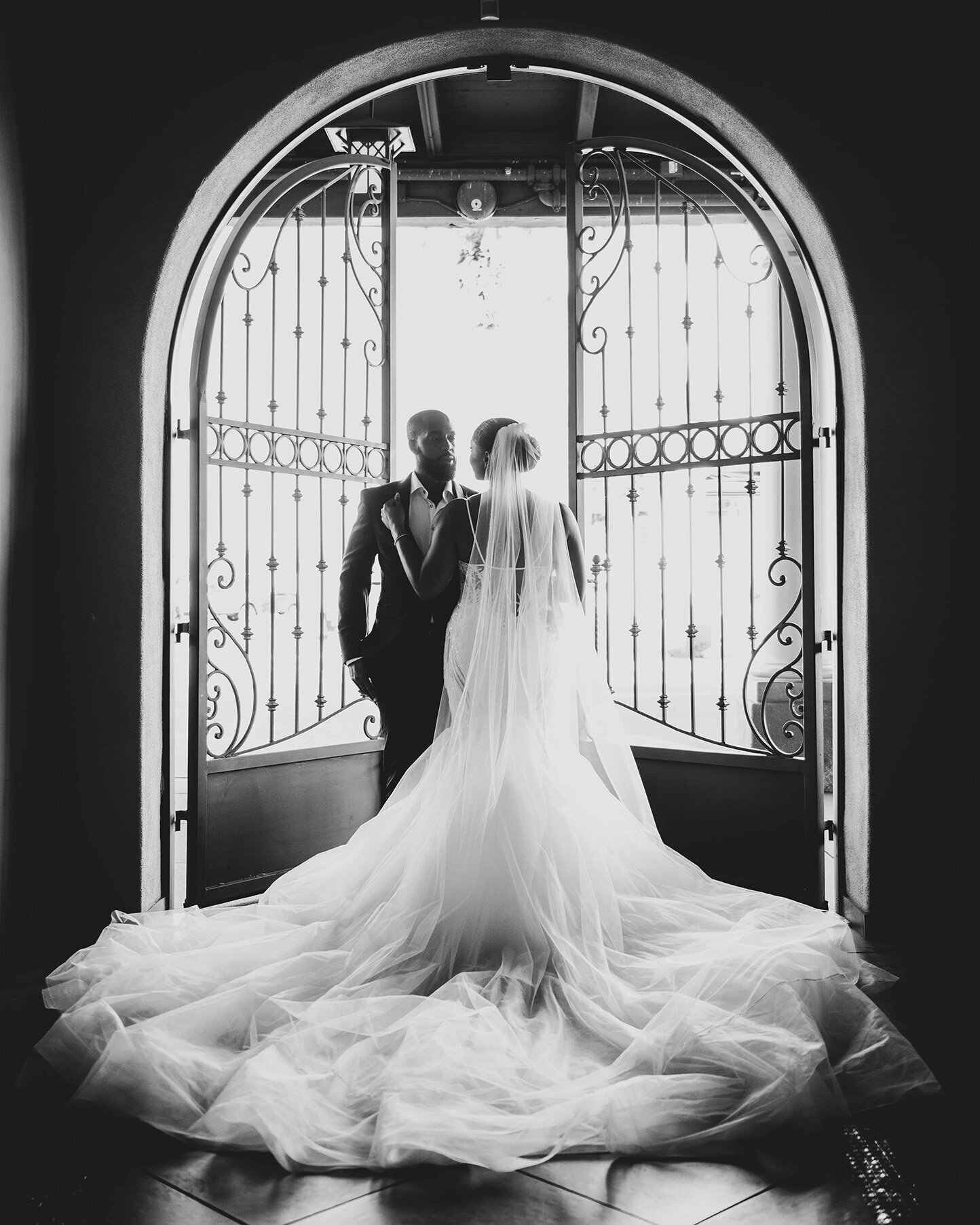 A bride and groom stand framed by the iconic arched double doors at SoHo63, a Chandler wedding and event venue. The bride’s voluminous gown and veil flow toward the camera in this black-and-white portrait, capturing the timeless elegance and architectural beauty of SoHo63.