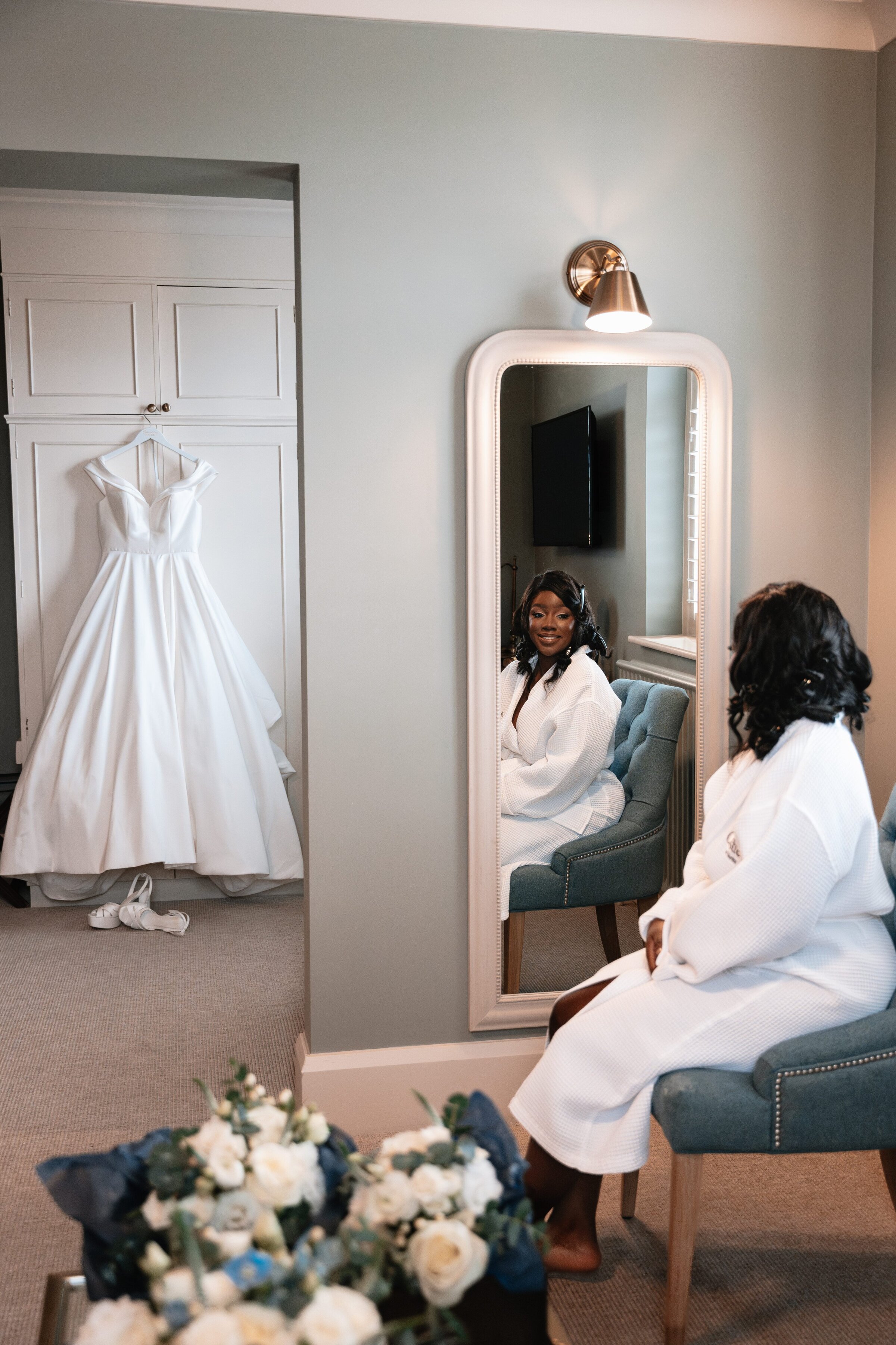 Bride sat in dressing gown looking in mirror, her wedding dress hangs in the background