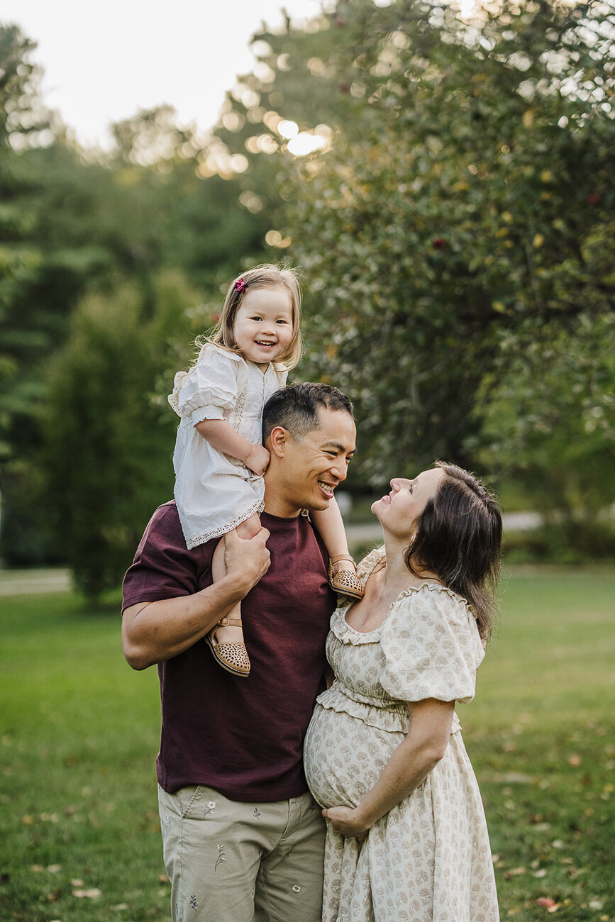 toddler girl sits on dads shoulders while he smiles at pregnant wife