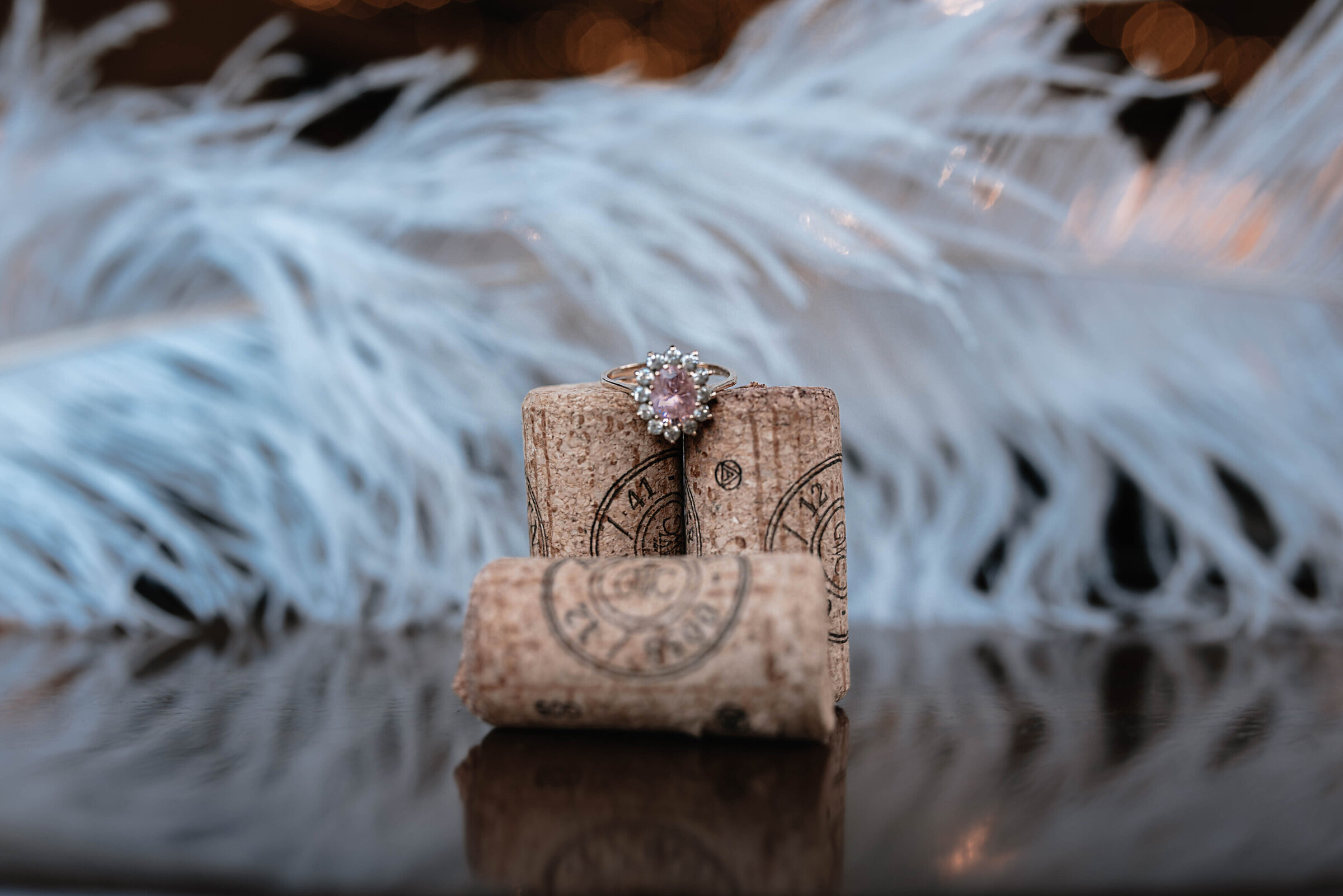 Rose and diamond engagement ring seated on top of champagne corks in front of white feathered backdrop