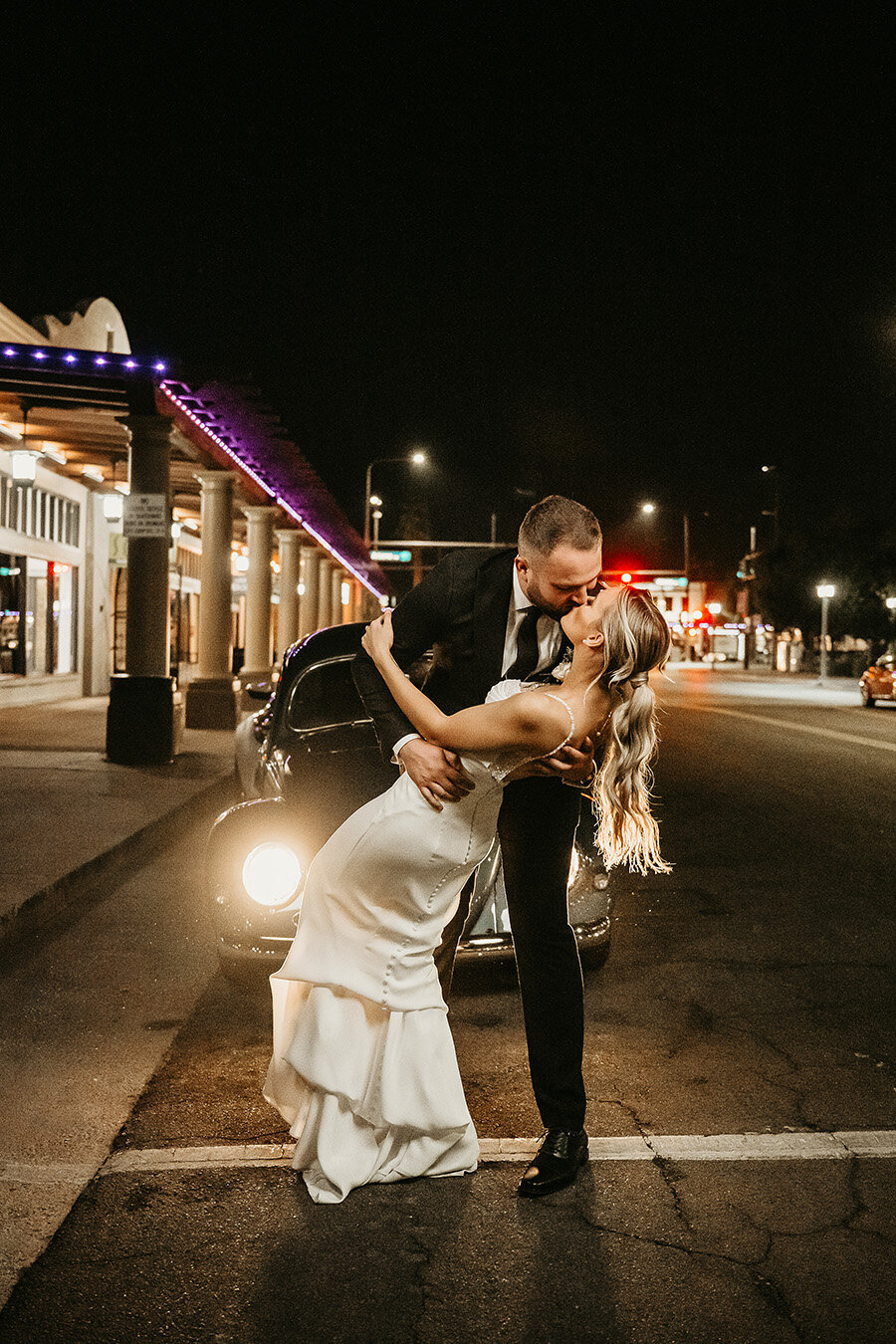 A groom dips his bride for a kiss under the city lights outside SoHo63, a Chandler wedding and event venue. With a vintage car behind them and the venue’s glowing purple lights lining the roof, the couple’s romantic night-time sendoff captures the charm and energy of downtown Chandler.