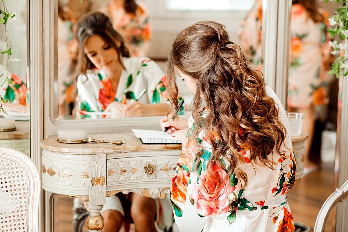 Bride sitting at a vintage desk in the bridal suite at Drakewood Farms writing a note to her husband to be while wearing an elegant silk robe with orange, ivory and green details