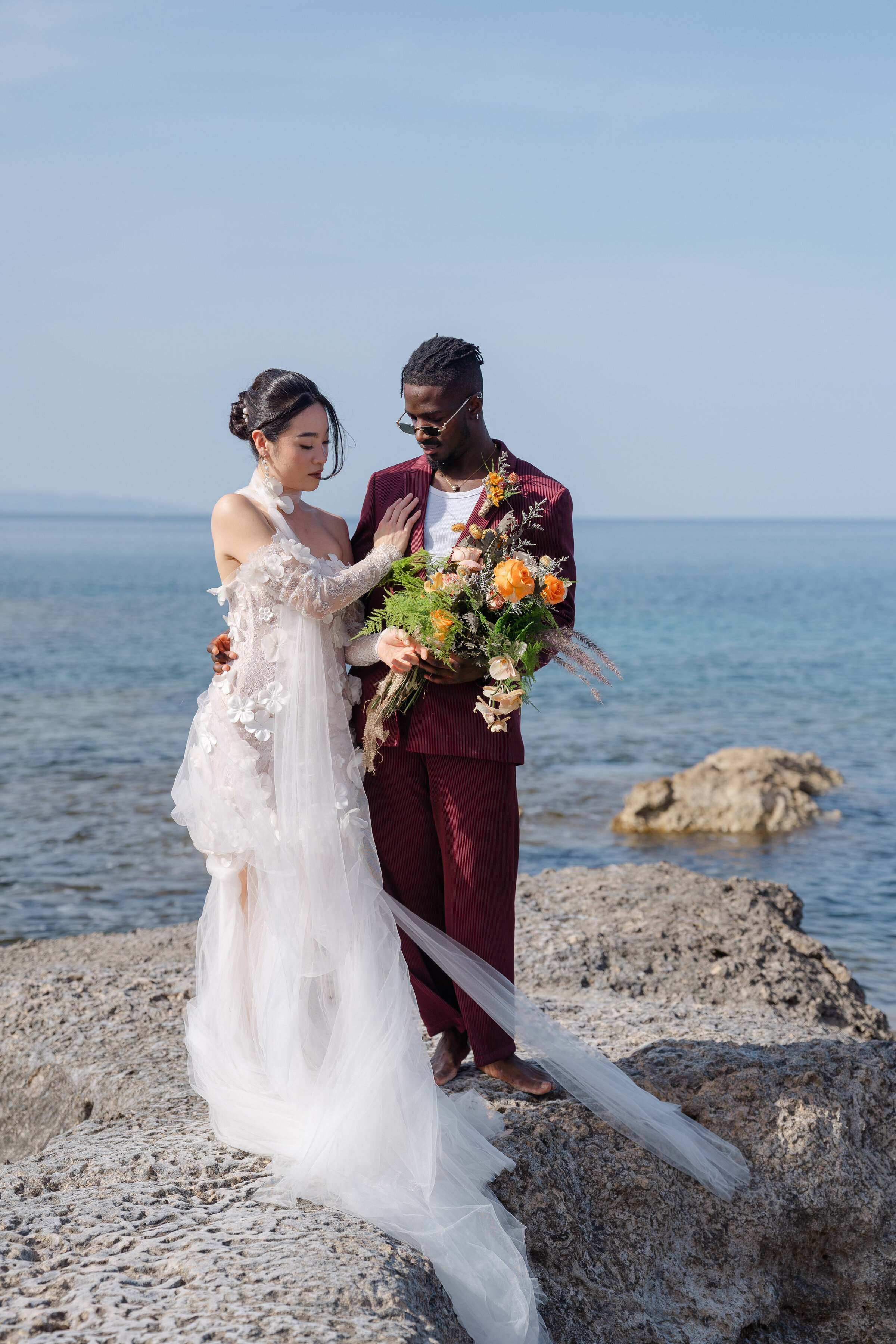 Bride and groom standing barefoot on seaside rocks in Milos, Greece, during a multicultural destination wedding shoot by Chi Ramos Photography.
