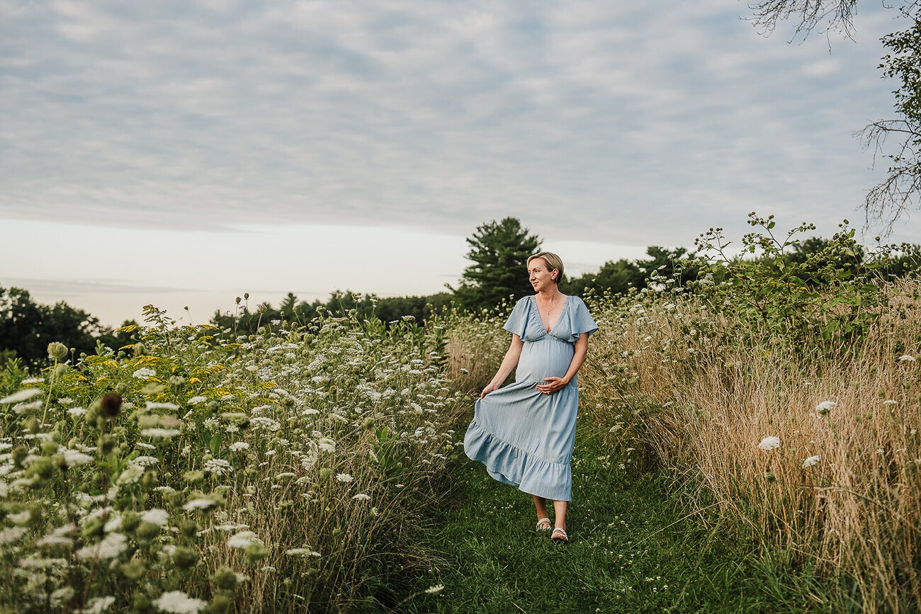 woman in blue dress walks through flower field for maternity photo session