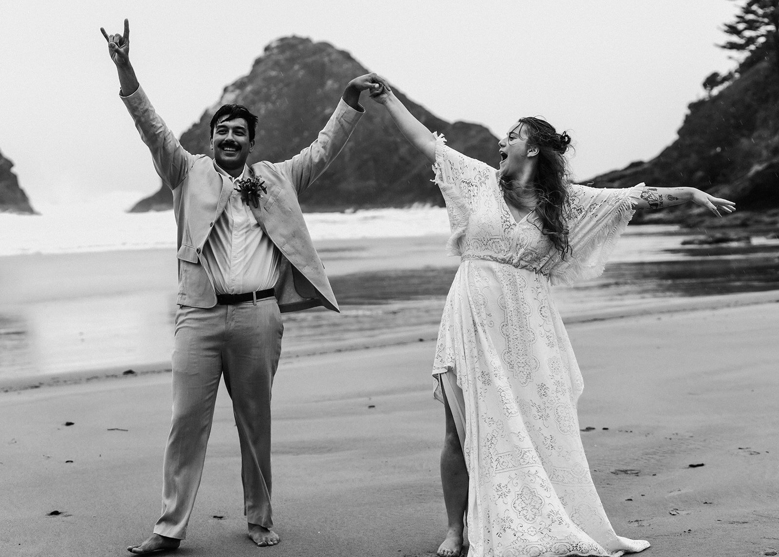 a couple in wedding attire cheer on a rocky beach after their adventure elopement ceremony. The brides smiles at her groom and the groom looks directly at their adventure elopement photographers as they cheer