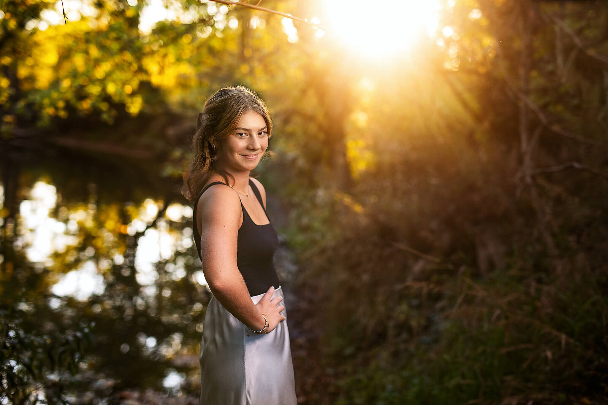 High school senior wearing silk skirt with blank tank top standing in front of a river and the sun setting behind her