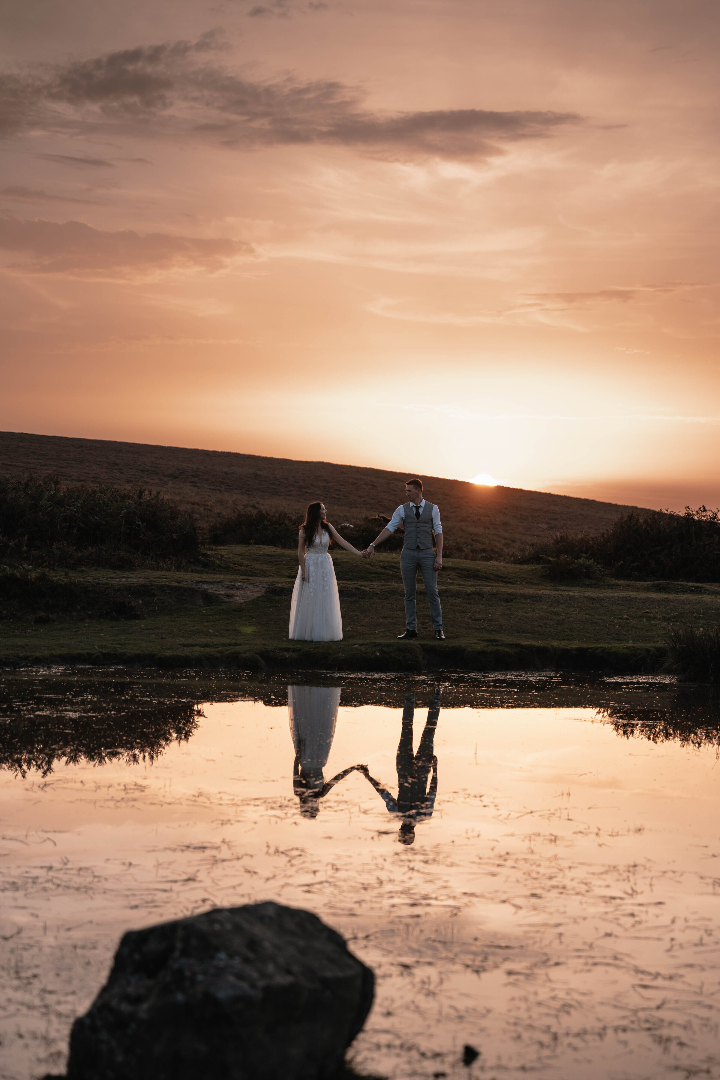 Bride and Groom in wedding attire holding hands in front of pond with golden sunset in the background