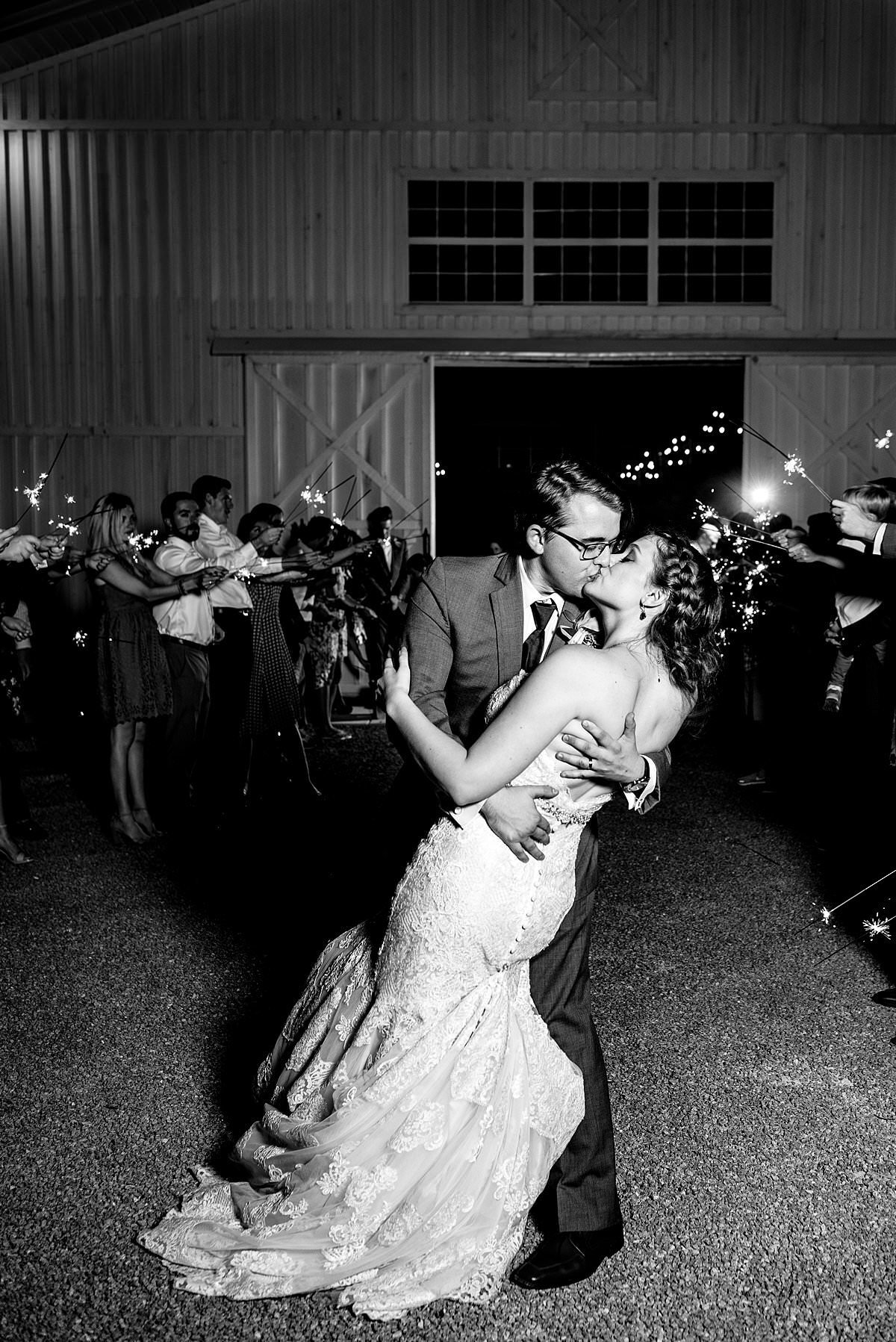Black and white photo of groom dipping bride and kissing her during their sparkler exit at White Dove Barn