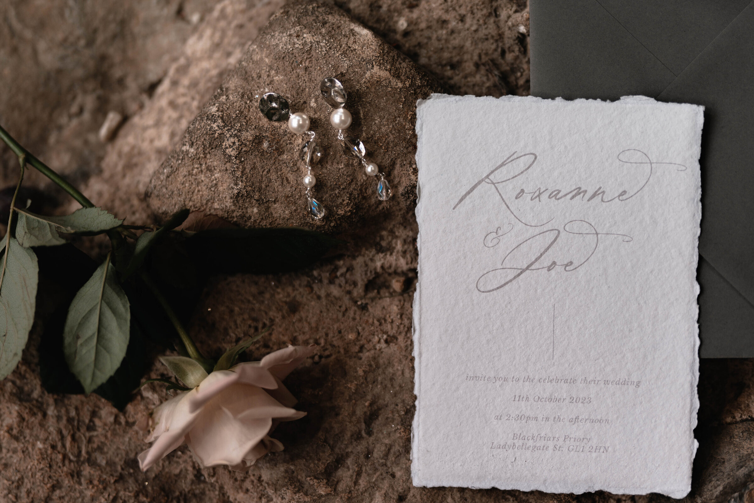 A wedding invitation, pear earrings and a single rose laid on top of a rocky surface