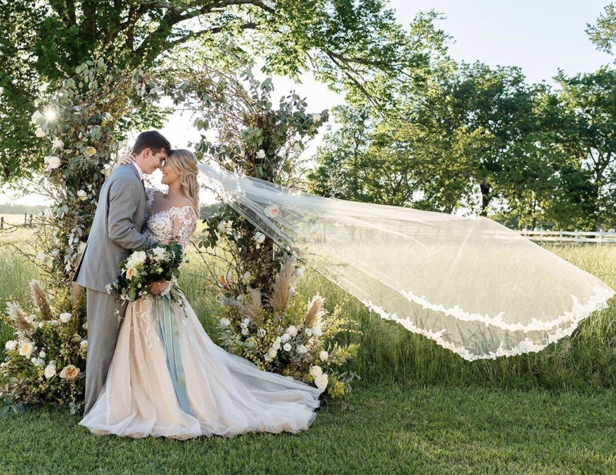 A bride and groom posing in a field in front of a gorgeous floral installation. The bride is facing the groom and her veil is flowing behind her