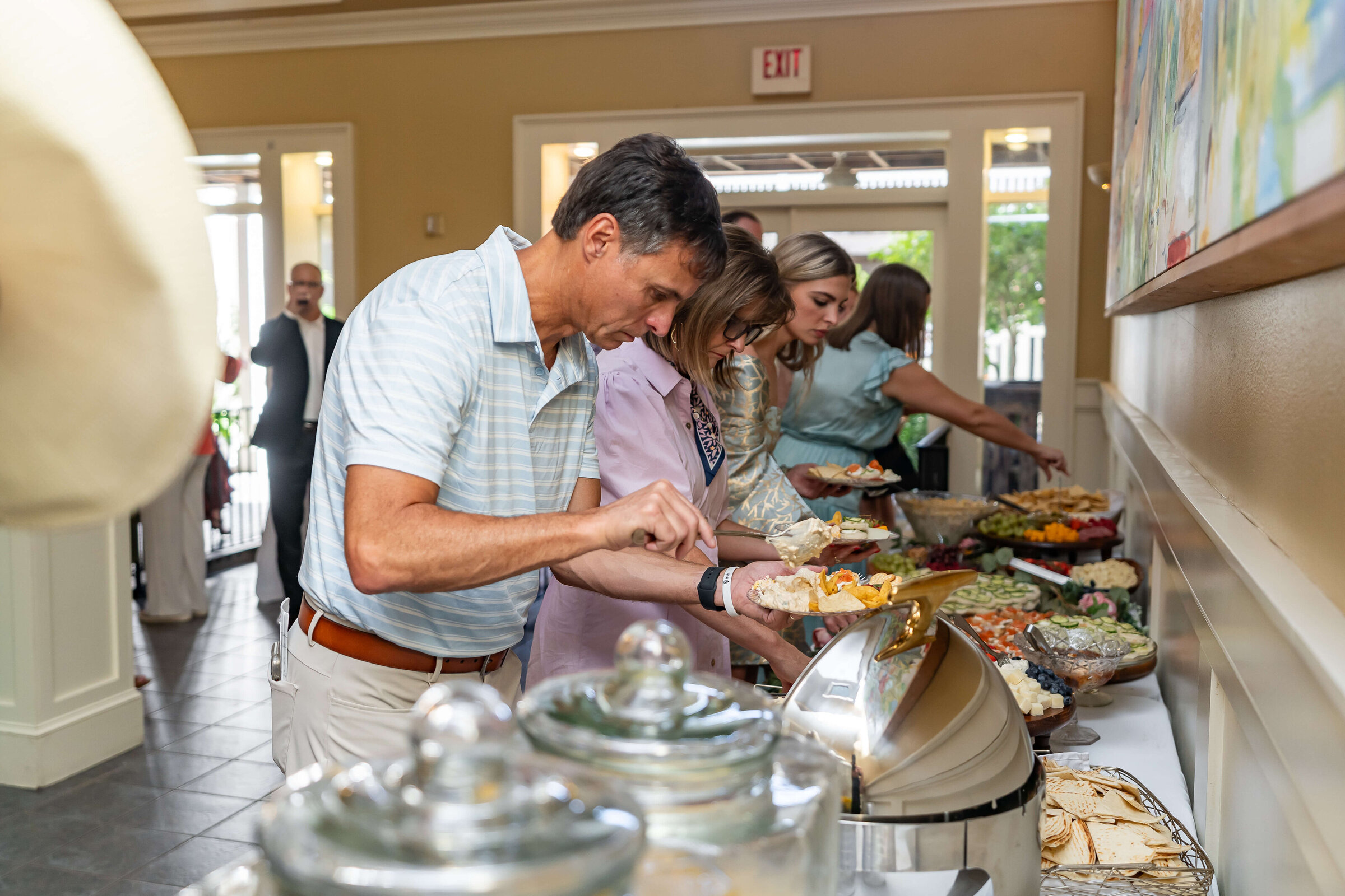 A group of people serve themselves food from a buffet table indoors, selecting from various dishes and salads. The setting appears casual and social, with natural light coming through the windows.