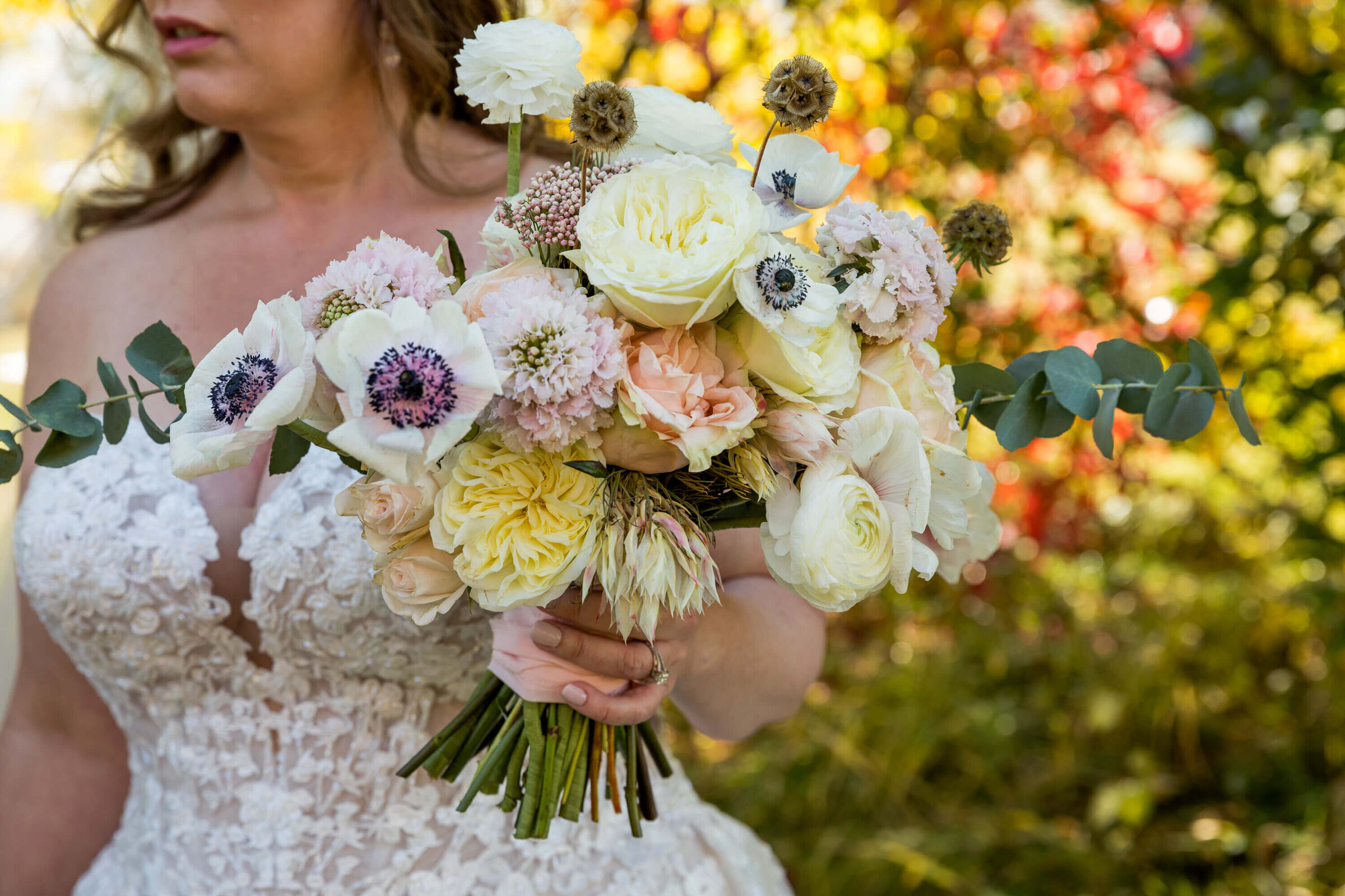 A close-up of the bride’s bouquet at Diamond Cross Ranch shows ivory garden roses, blush spray roses, white anemones with dark centers, and eucalyptus branches. The soft, romantic blooms contrast with the vibrant autumn leaves in the background.