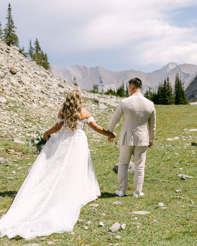 An adventurous elopement photograph of a bride and groom holding hands as they walk into the vast wilderness after their private helicopter elopement at Mount Charles Stewart in Banff National Park. The bride elegantly plays with her dress as towering mountains surround them, creating a breathtaking and timeless moment of love, freedom, and untouched beauty.