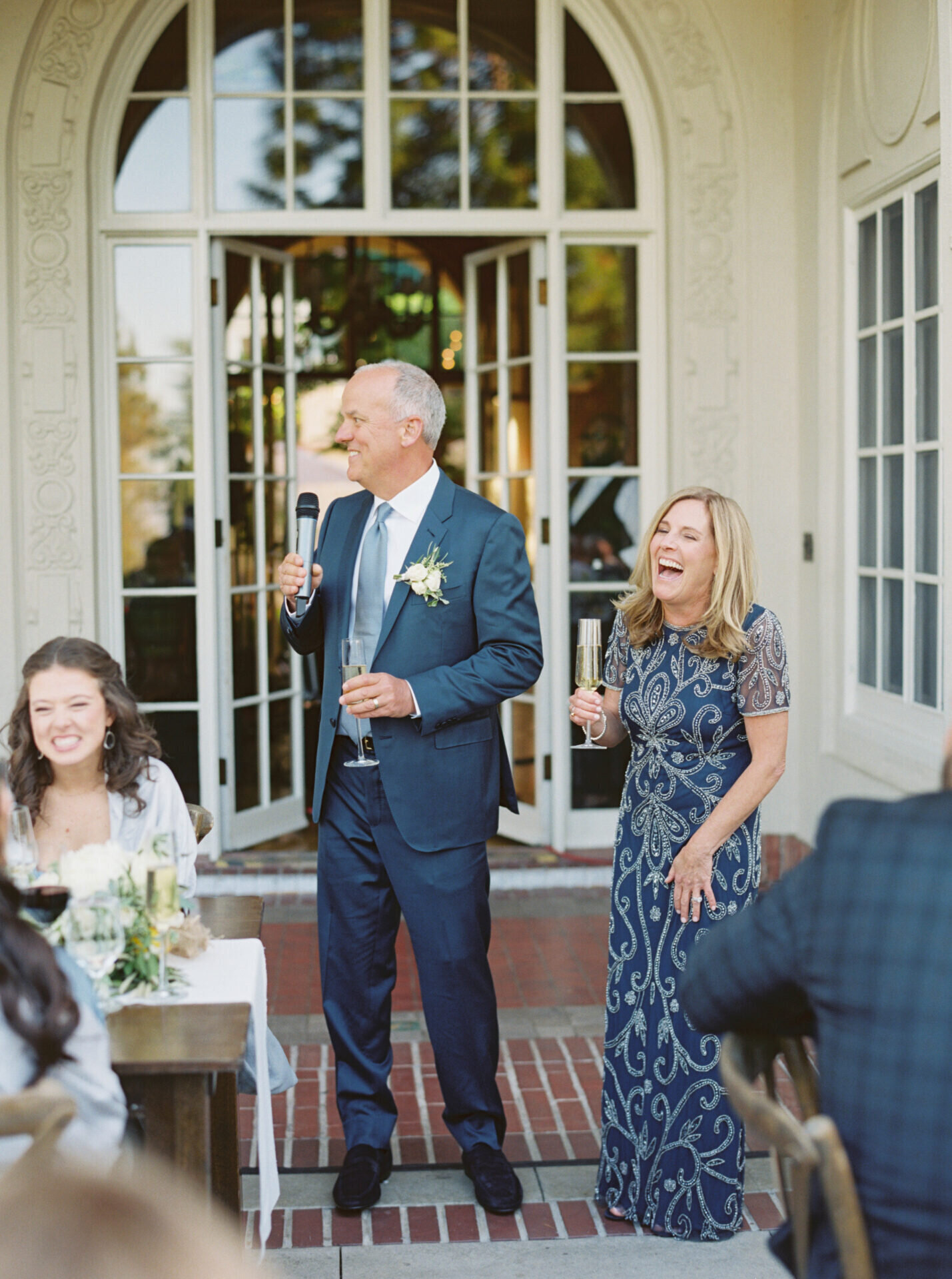 parents of the bride in navy blue laughing giving a toast at the wedding reception