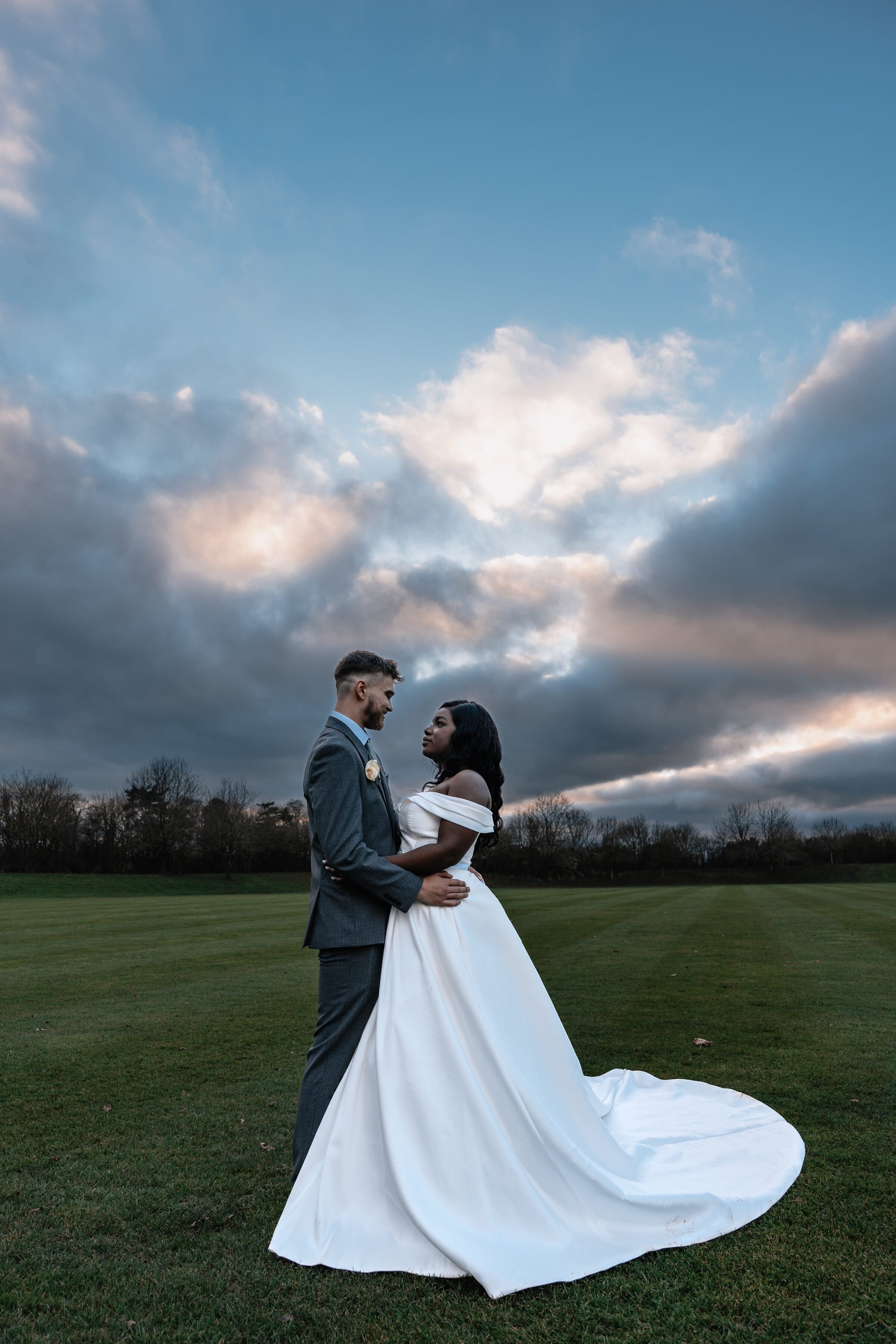 Bride and Groom in wedding attire stood close together in a field with a moody sunset sky behind them