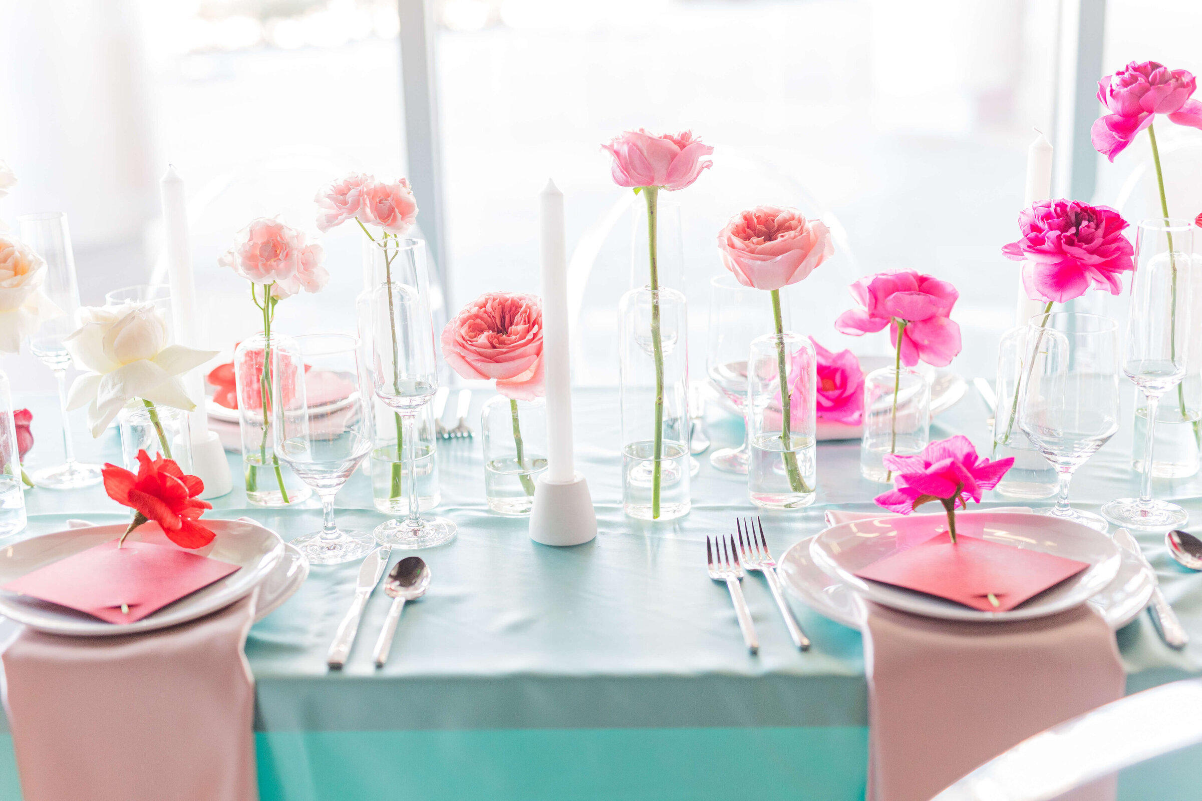 A table with a teal tablecloth, plates, pink napkins, and roses all in individual vases