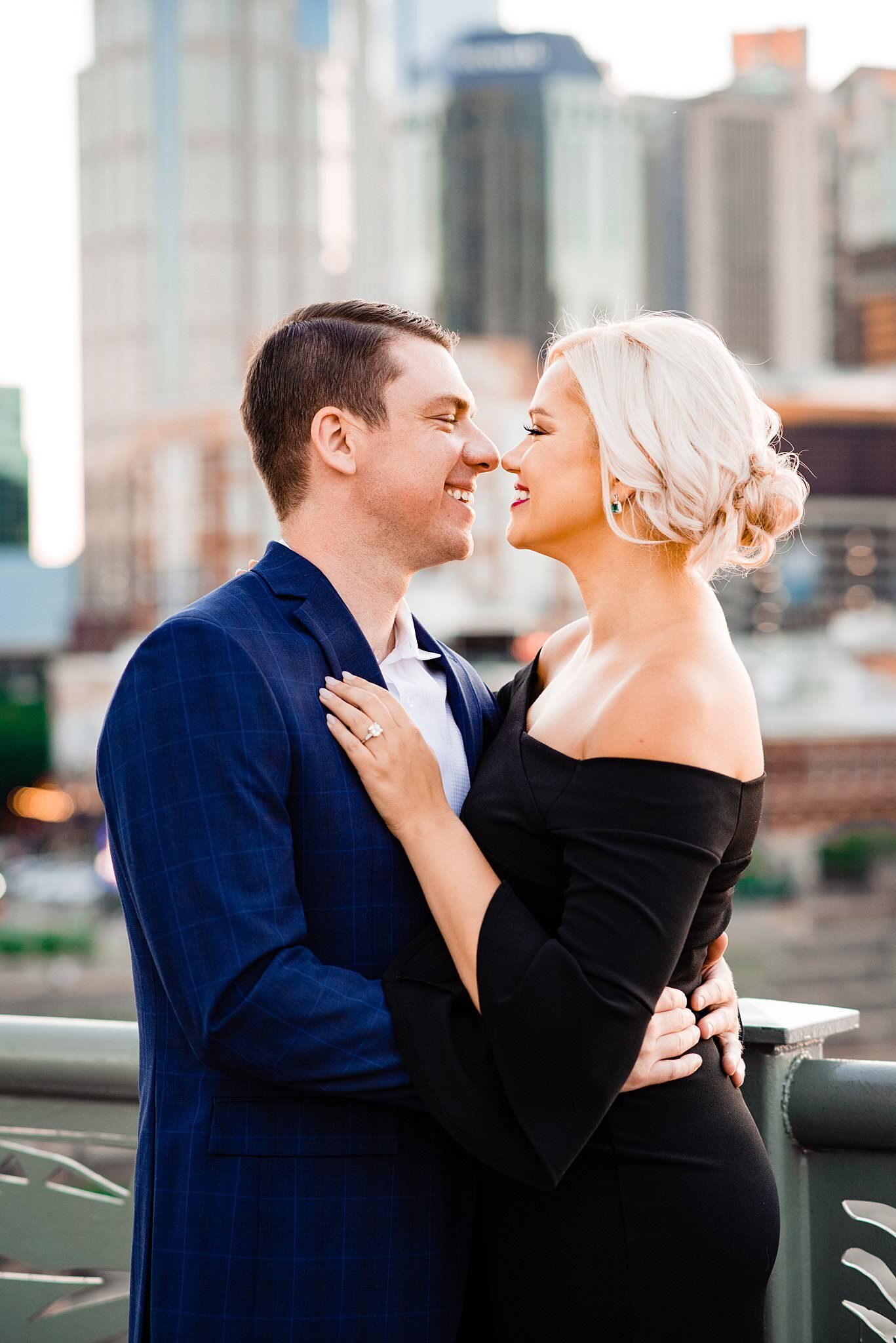 Close up photo of couple dressed elegantly is black and blue standing on the pedestrian bridge in Nashville with the city behind them