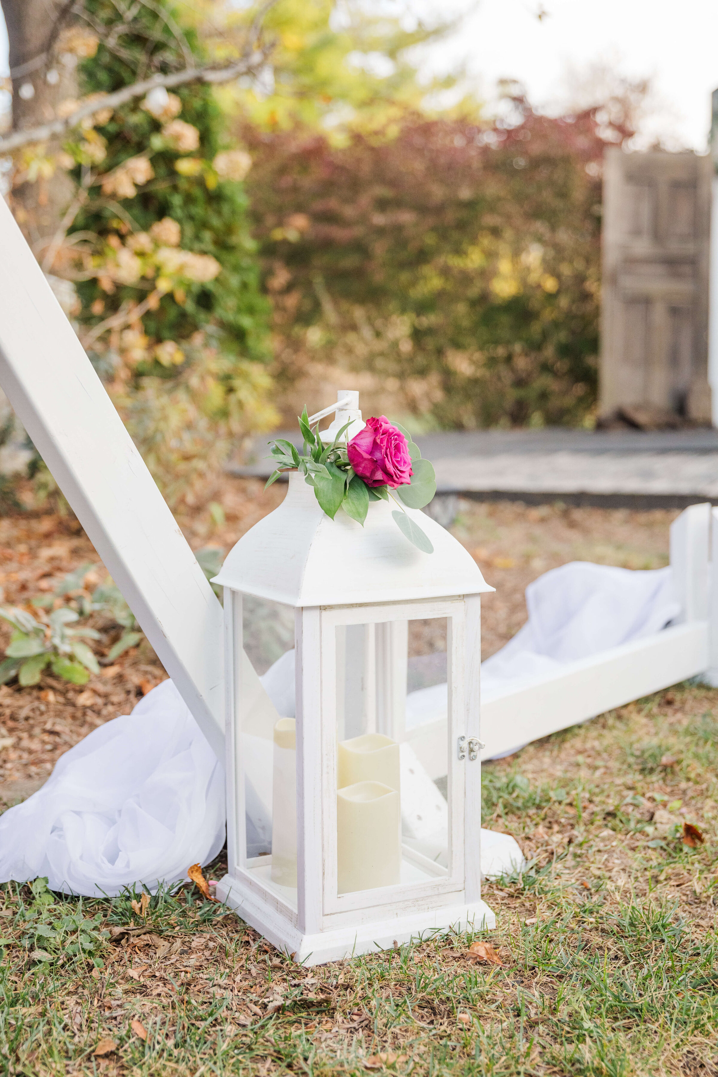 A photo of a white wedding lantern  on the grass by a white arch. The lantern has pink and white roses on top of it