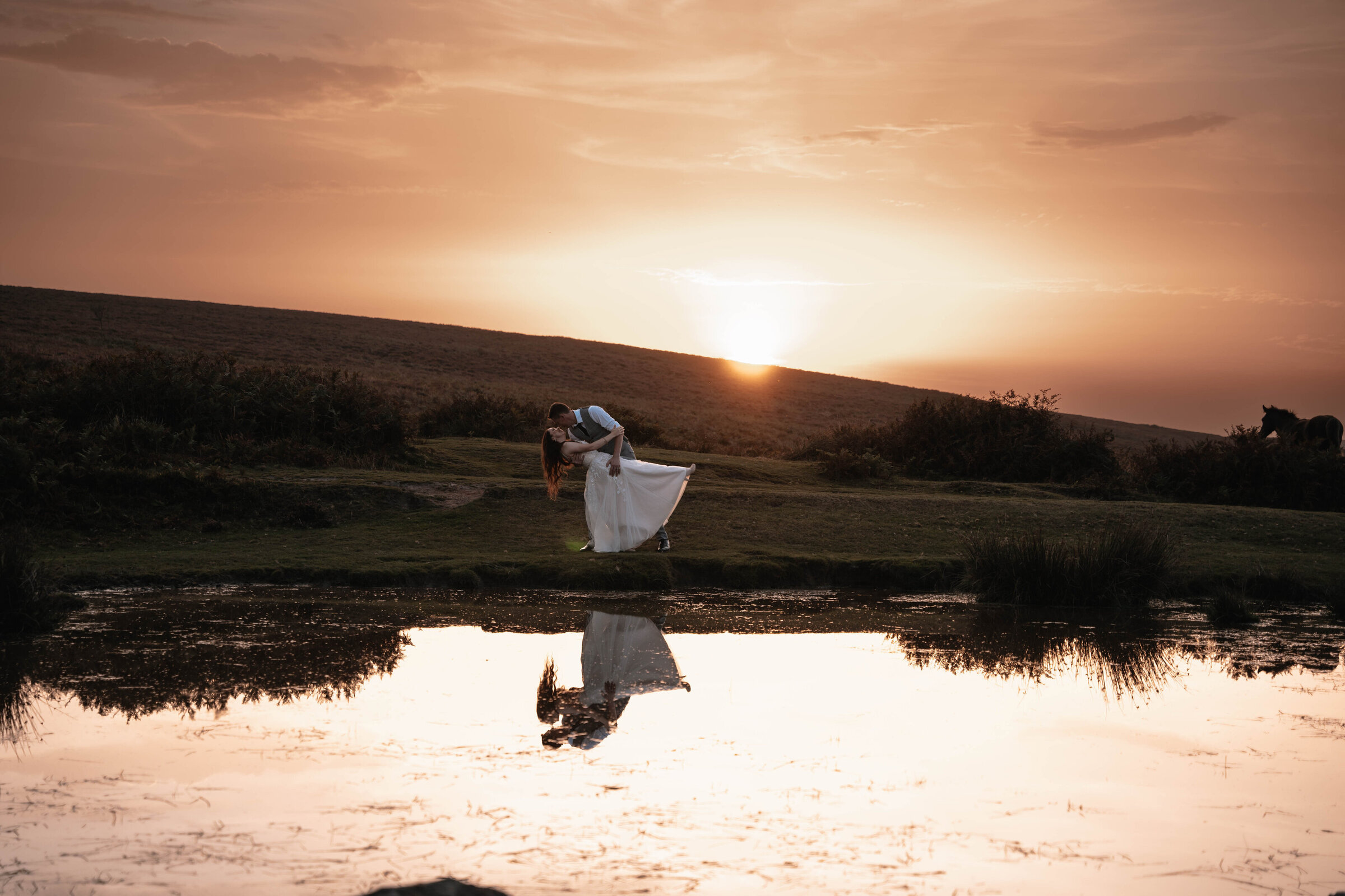 Bride and Groom in wedding attire having a dip kiss in front of a pond with a golden sunset in the background
