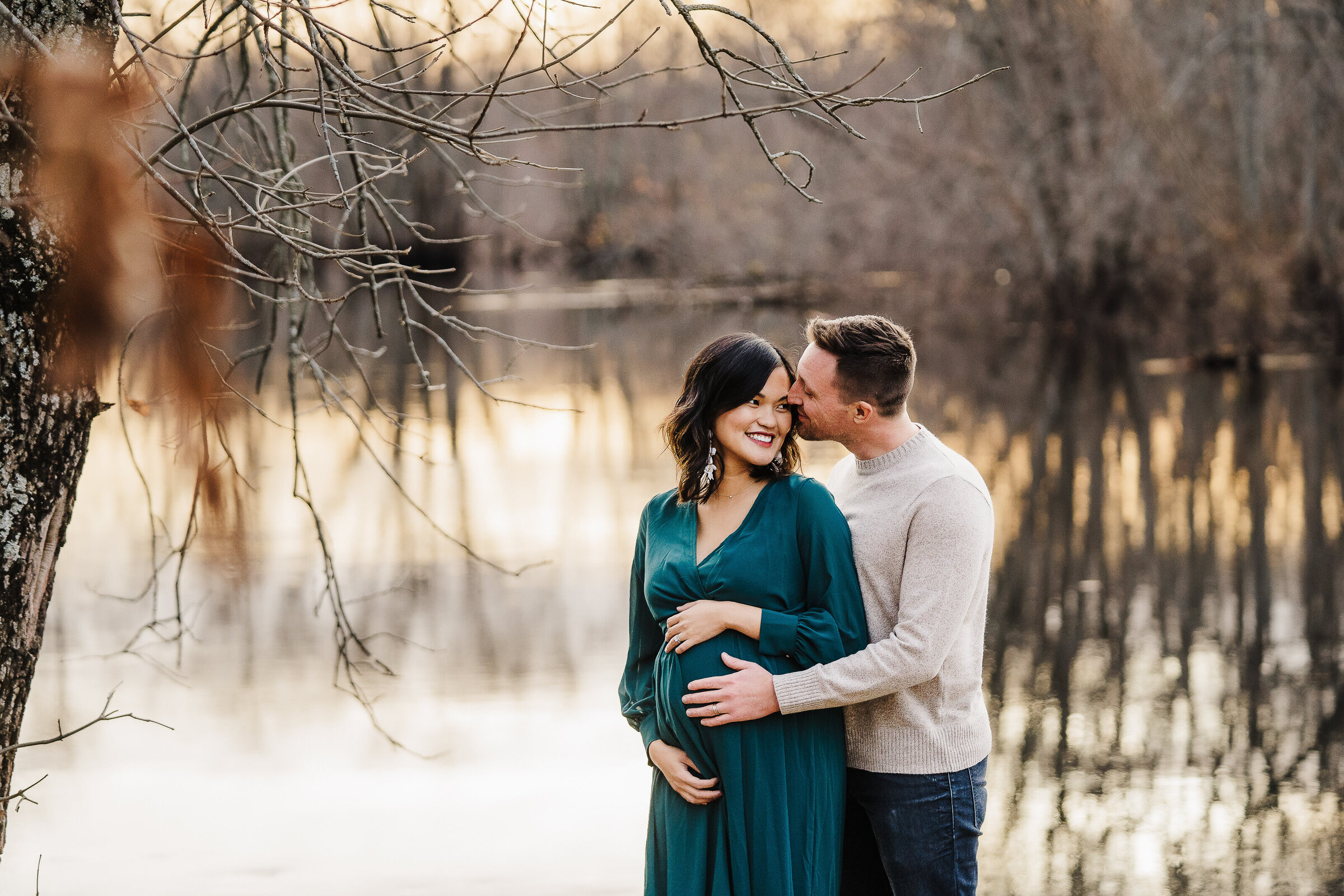 pregnant couple stands in front of a lake at sunset