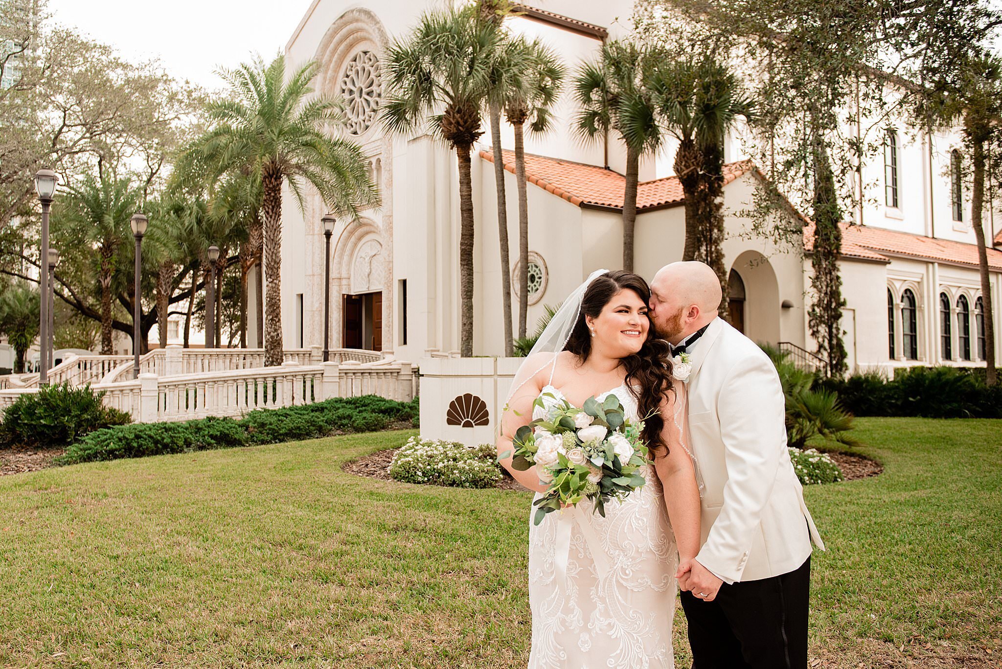 Groom whispering in the ear of his wife while standing before a large Catholic church in downtown central Orlando