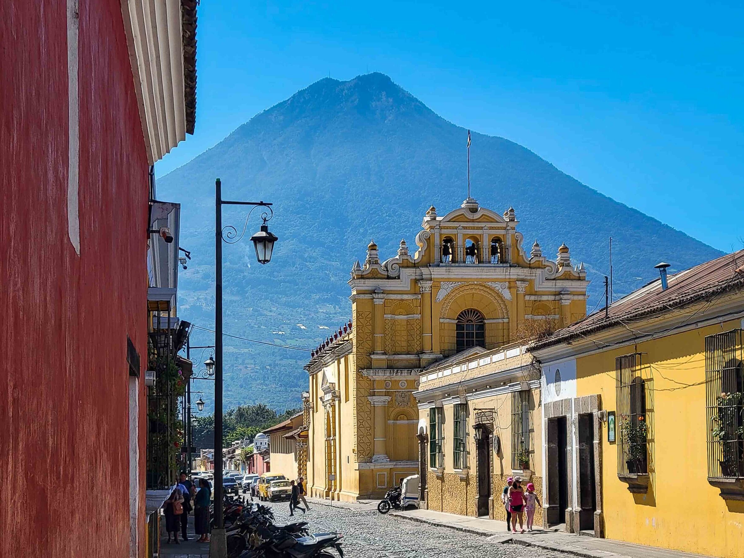 A street scene in Antigua, Guatemala showing a yellow church and a red wall with a volcano behind