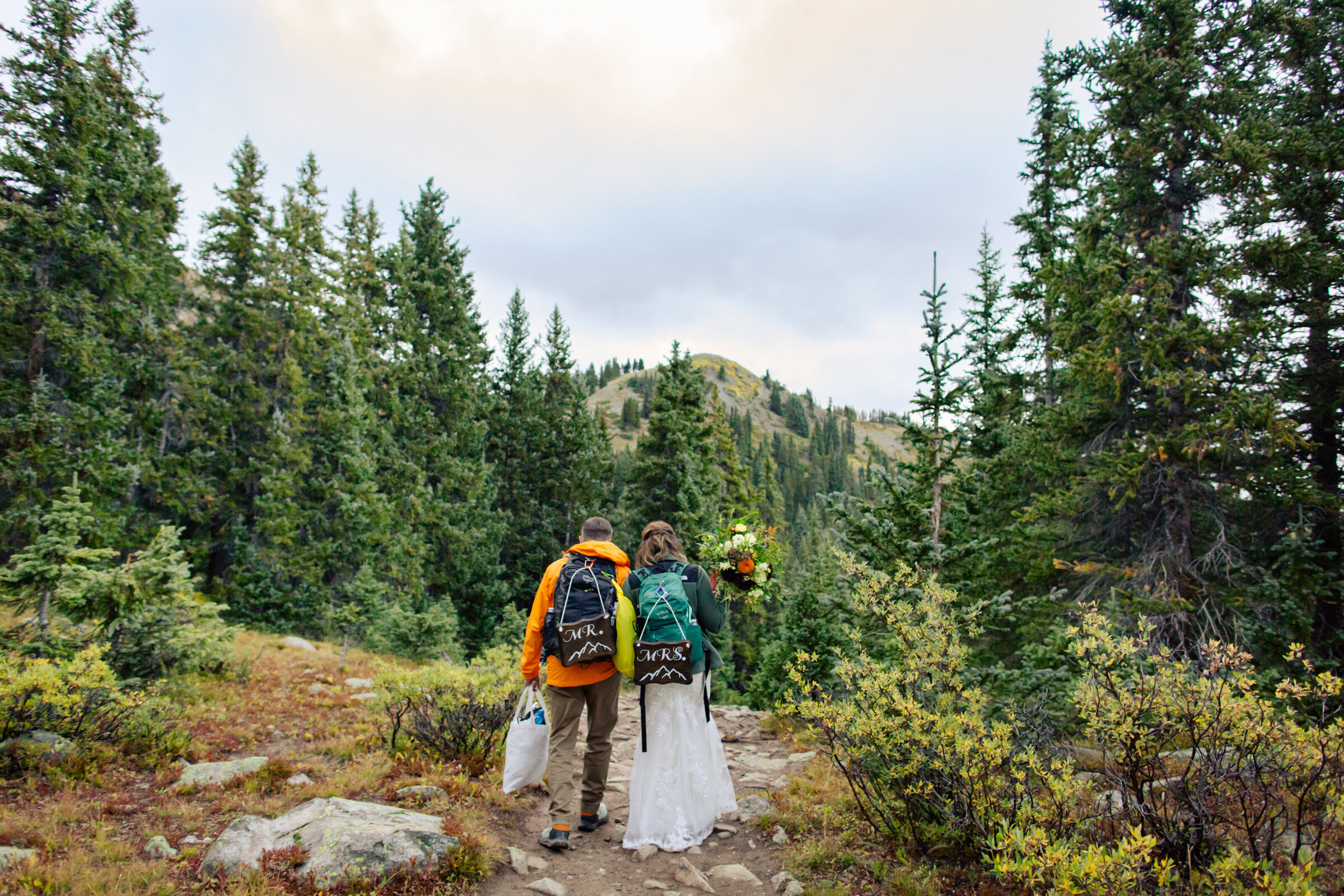 Bride and groom hike along a trail for their elopement while wearing "Mr. and Mrs. signs on their backpacks"