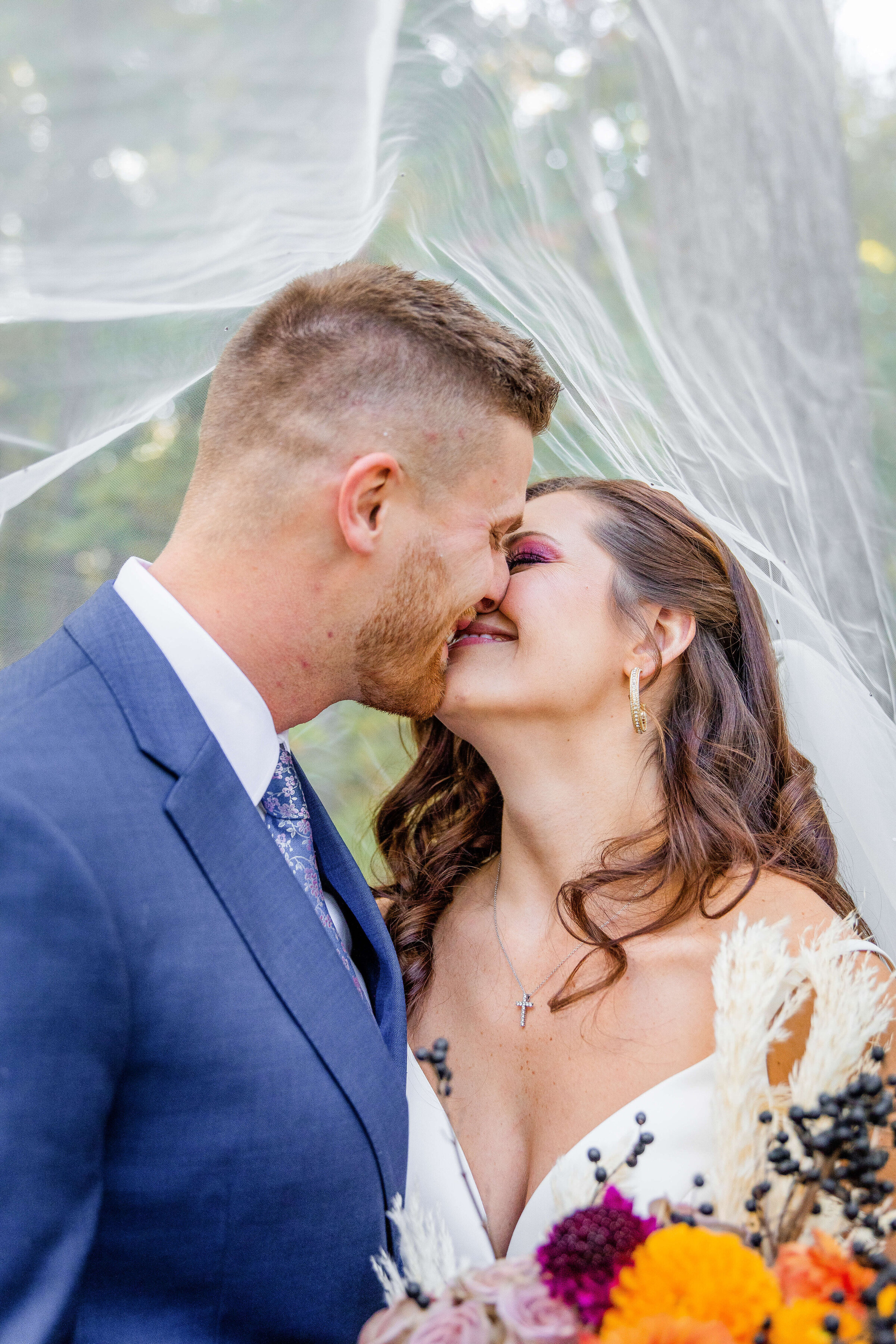 A bride and groom laugh while they kiss and she has her veil over their heads. She is holding a fall colors inspired bouquet