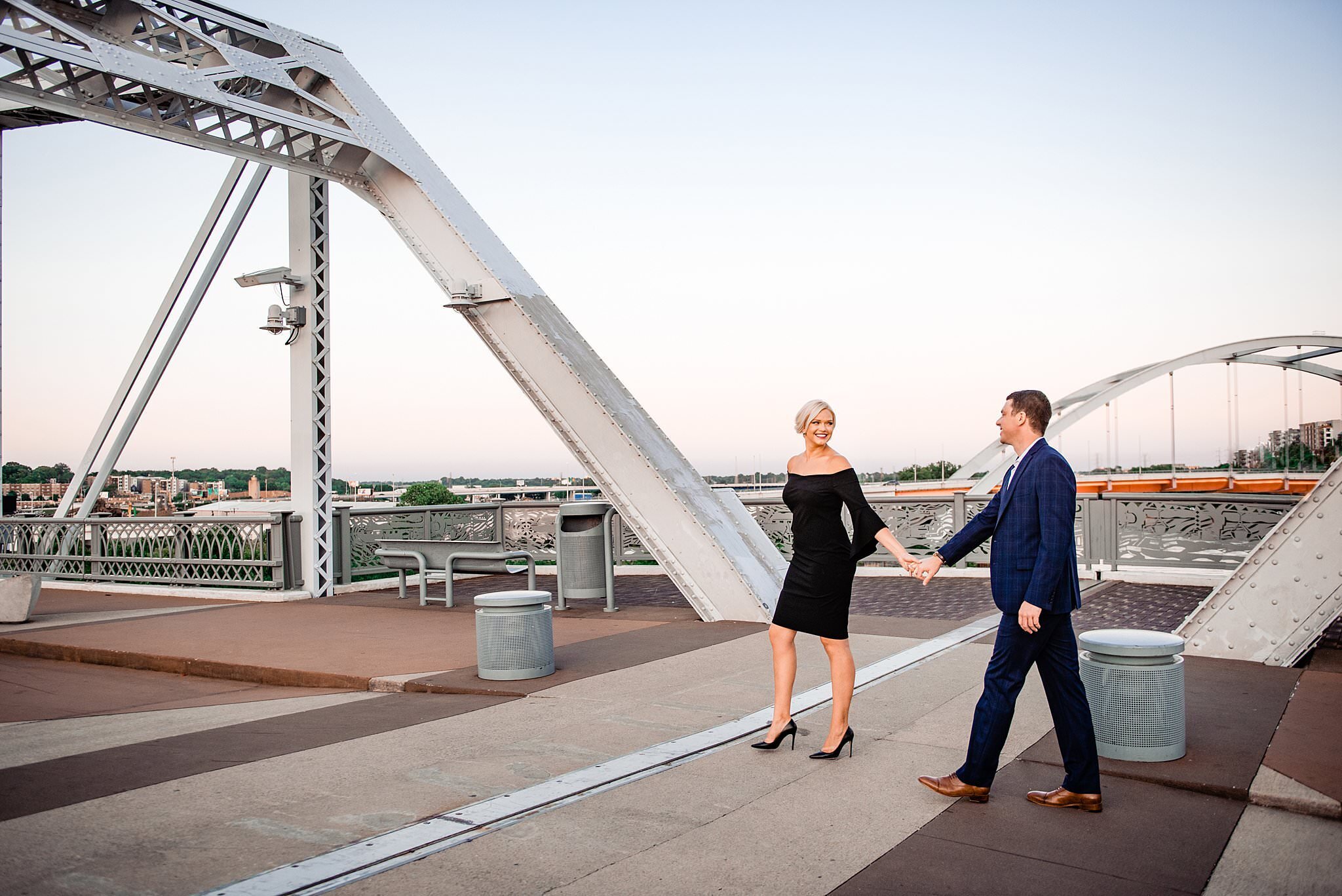 Girl wearing an elegant knee length black dress leading her fiance across the Nashville pedestrian bridge, he's in a navy suit
