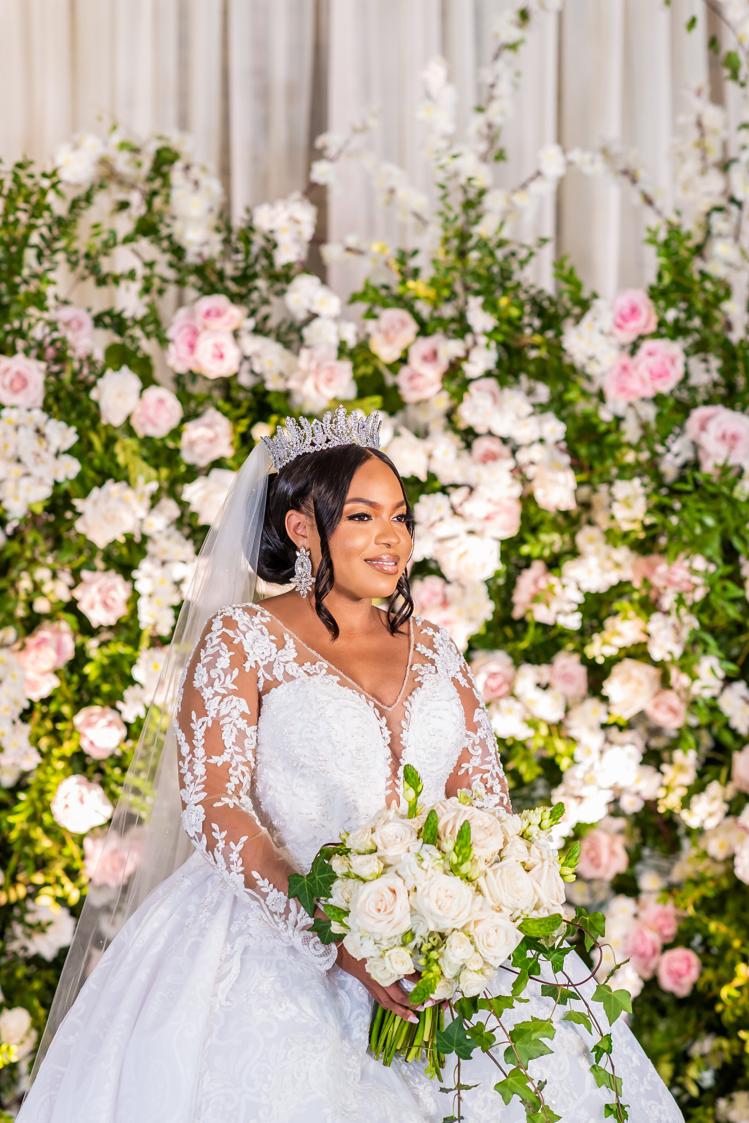 Bridal portrait in lace gown and tiara holding a white rose bouquet in front of lush floral wall at a romantic wedding ceremony in Birmingham, AL by Wells Design Co.