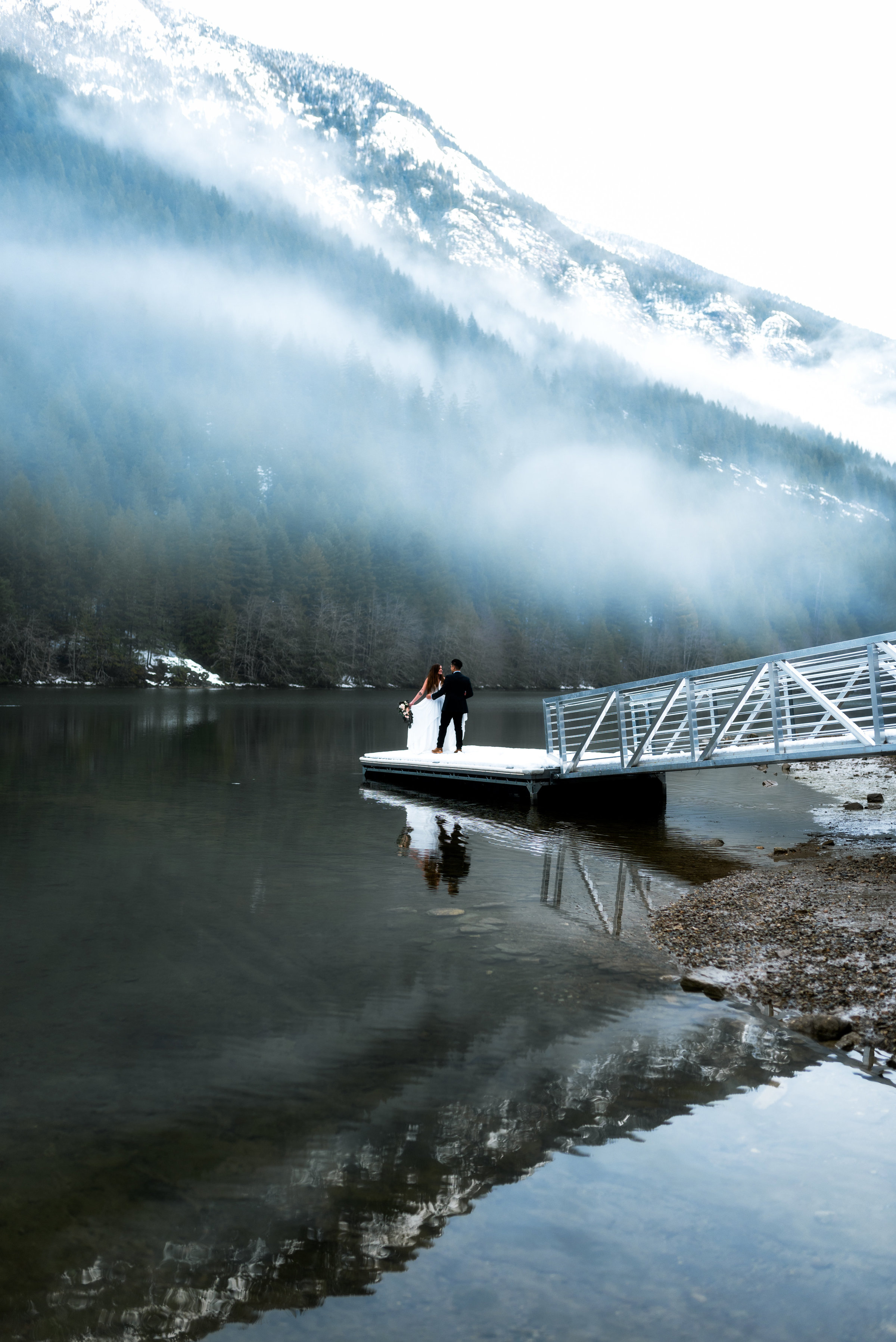 Wide angle shot of a couple in wedding attire on a dock looking out at snowy lake