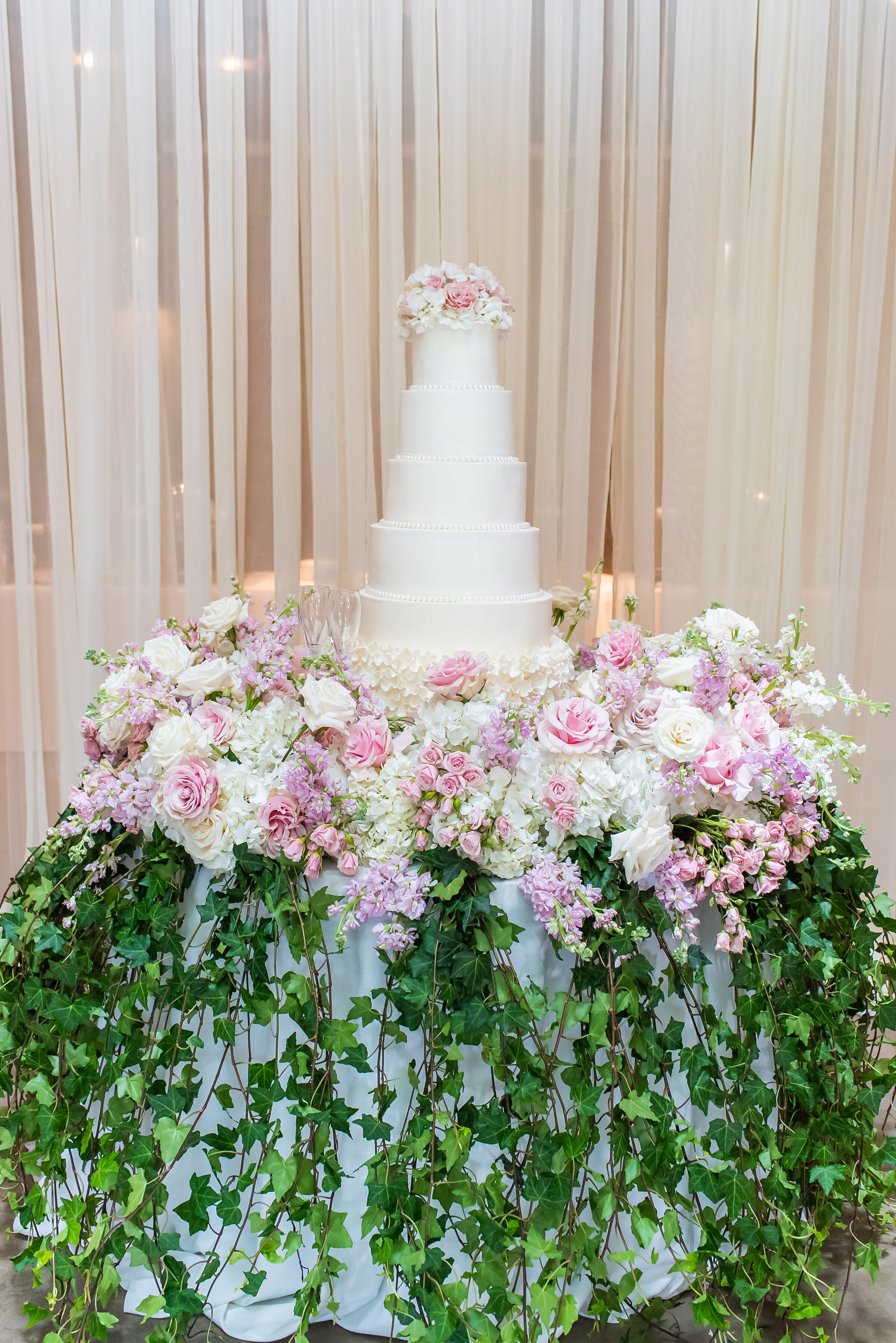 Elegant five-tier white wedding cake surrounded by lush pink, white, and lavender florals with cascading greenery, designed by Wells Design Co for a luxury wedding reception in Birmingham, AL