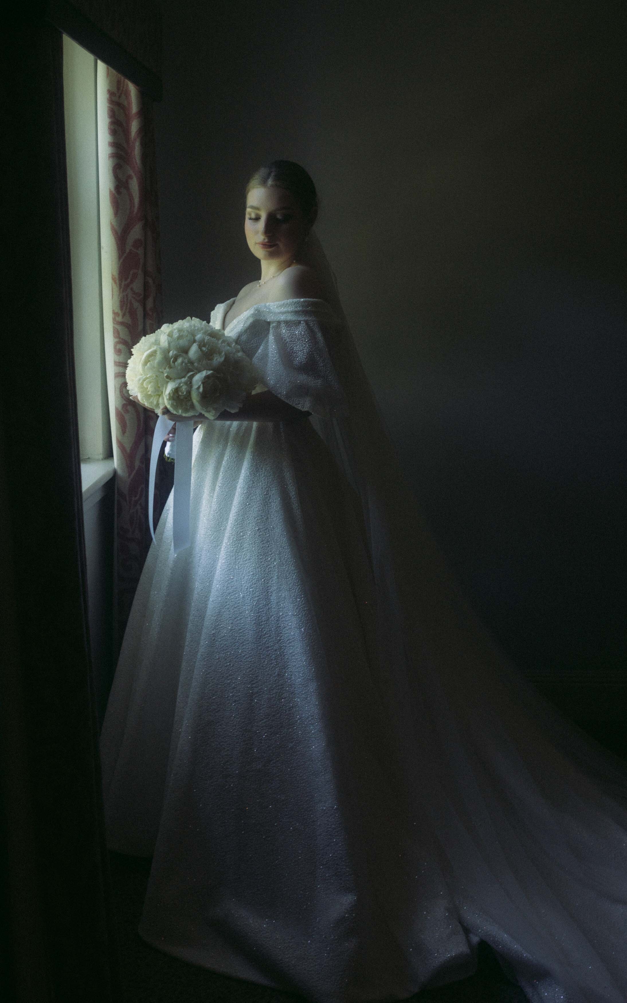 A stunning modern bride posing gracefully with a bouquet of peonies next to a window, capturing her elegance and beauty on her wedding day