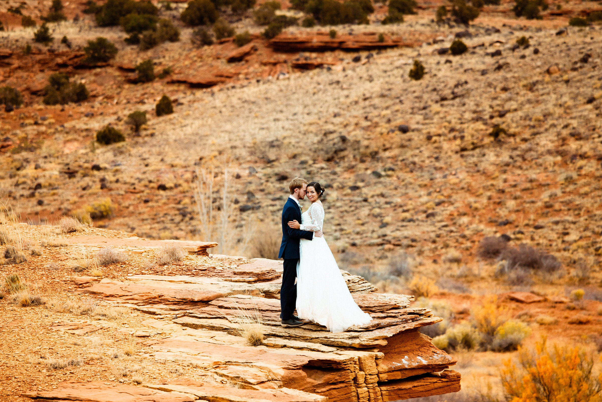 Bride and groom stand on a red rock cliff in Capitol Reef National Park near Moab, Utah holding each other closely. The bride is looking at the camera.